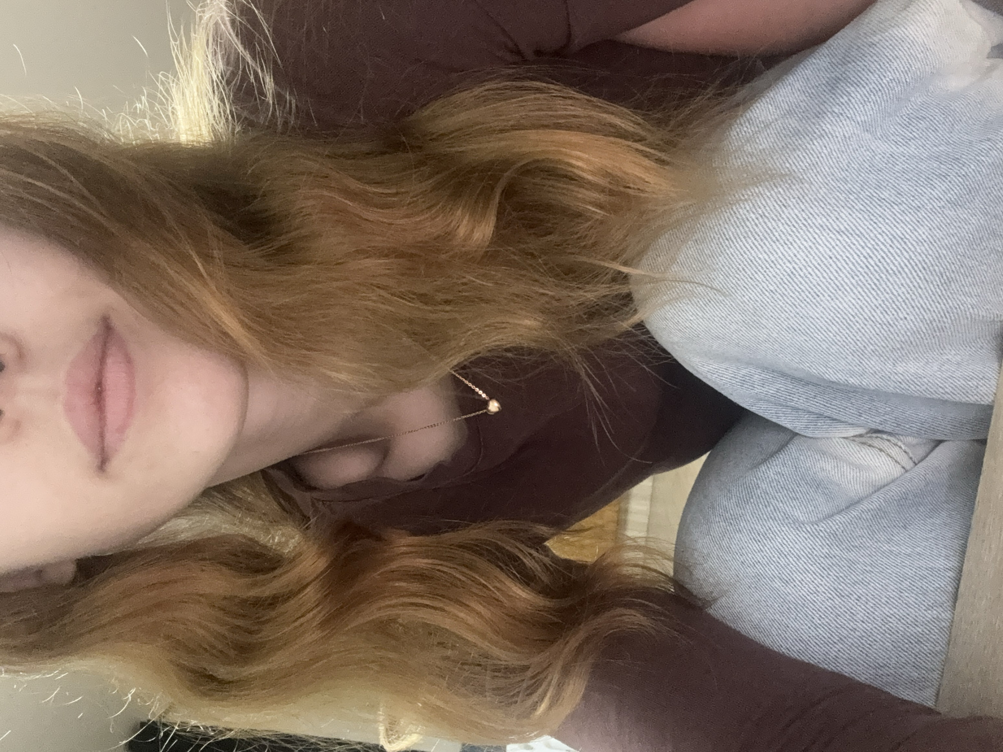 A person with long, wavy, reddish-blonde hair is shown from the chest up, crouching with their legs crossed. They are wearing a dark brown long-sleeved shirt and light blue jeans. A delicate gold necklace with a small heart pendant is visible around their neck. The background is blurred and appears to be an indoor setting with some light filtering from the left. No specific location in Gwangju, South Korea, can be identified from the image.