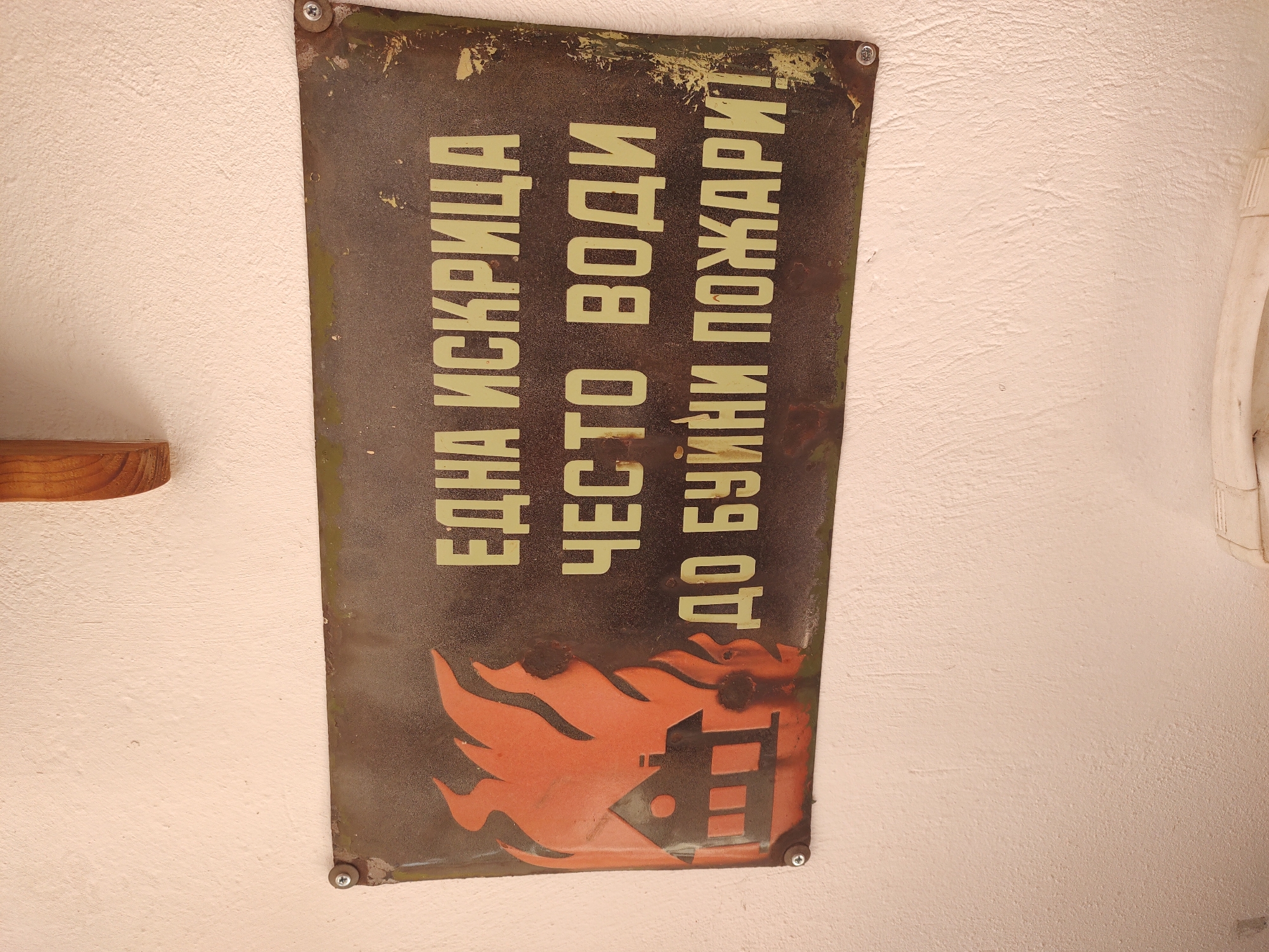 A weathered, rectangular metal sign is mounted on a textured, off-white wall. The sign has a dark background with lighter text and an image. To the left of the text, a stylized orange house engulfed in flames is depicted. The text, in a light yellow, reads: "ЕДНА ИСКРИЦА ЧЕСТО ВОДИ ДО БУЙНИ ПОЖАРИ!" which translates to "ONE SPARK OFTEN LEADS TO WILDFIRE!". The sign appears to be old and shows signs of rust and wear. It is attached to the wall with at least four screws. A small portion of a wooden shelf is visible at the top of the image. The overall setting appears to be indoors, possibly in a public building or common area. The location of the sign in Tervel, Bulgaria, is not discernable from the image itself.