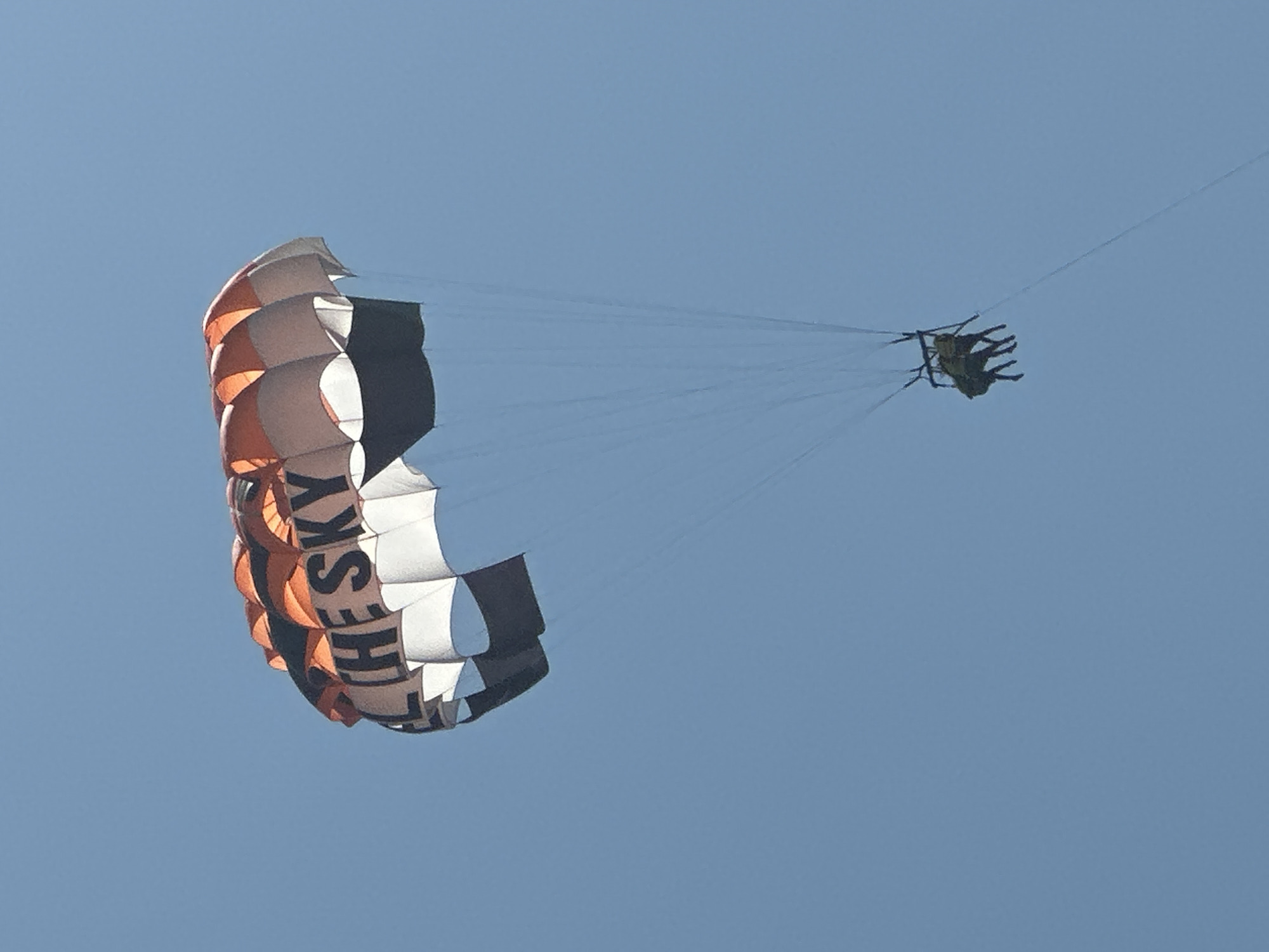 A parasail with the words "ALL THE SKY" printed on it is in flight against a clear blue sky. The parasail is multicolored, with sections of orange, white, and dark brown. Two people are suspended beneath the parasail in a harness. They appear to be wearing dark clothing. The scene is likely taking place over a body of water, as parasailing is a common water sport. The city of Boreti, Montenegro, is known for its coastal tourism and water activities, making it a plausible location for this image.