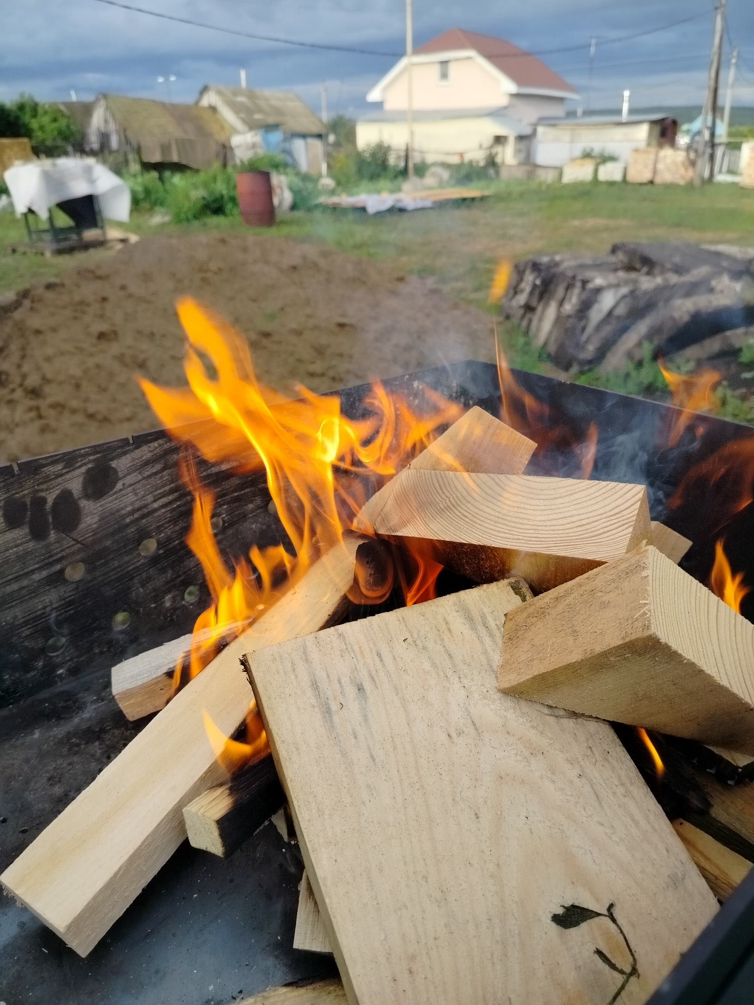 The image depicts a close-up of a metal grill filled with wood and flames. The wood pieces are light-colored and vary in size and shape, some stacked and some at an angle. Bright orange flames are actively burning among the wood, with wisps of smoke visible. The grill itself appears to be made of dark metal with a patterned texture. In the background, out of focus, a rural landscape is visible. This includes dirt or sand, green grass, various structures resembling houses or sheds with weathered roofs, and a cloudy sky. The overall scene suggests an outdoor activity, possibly a barbecue or bonfire, in a semi-rural or village setting.