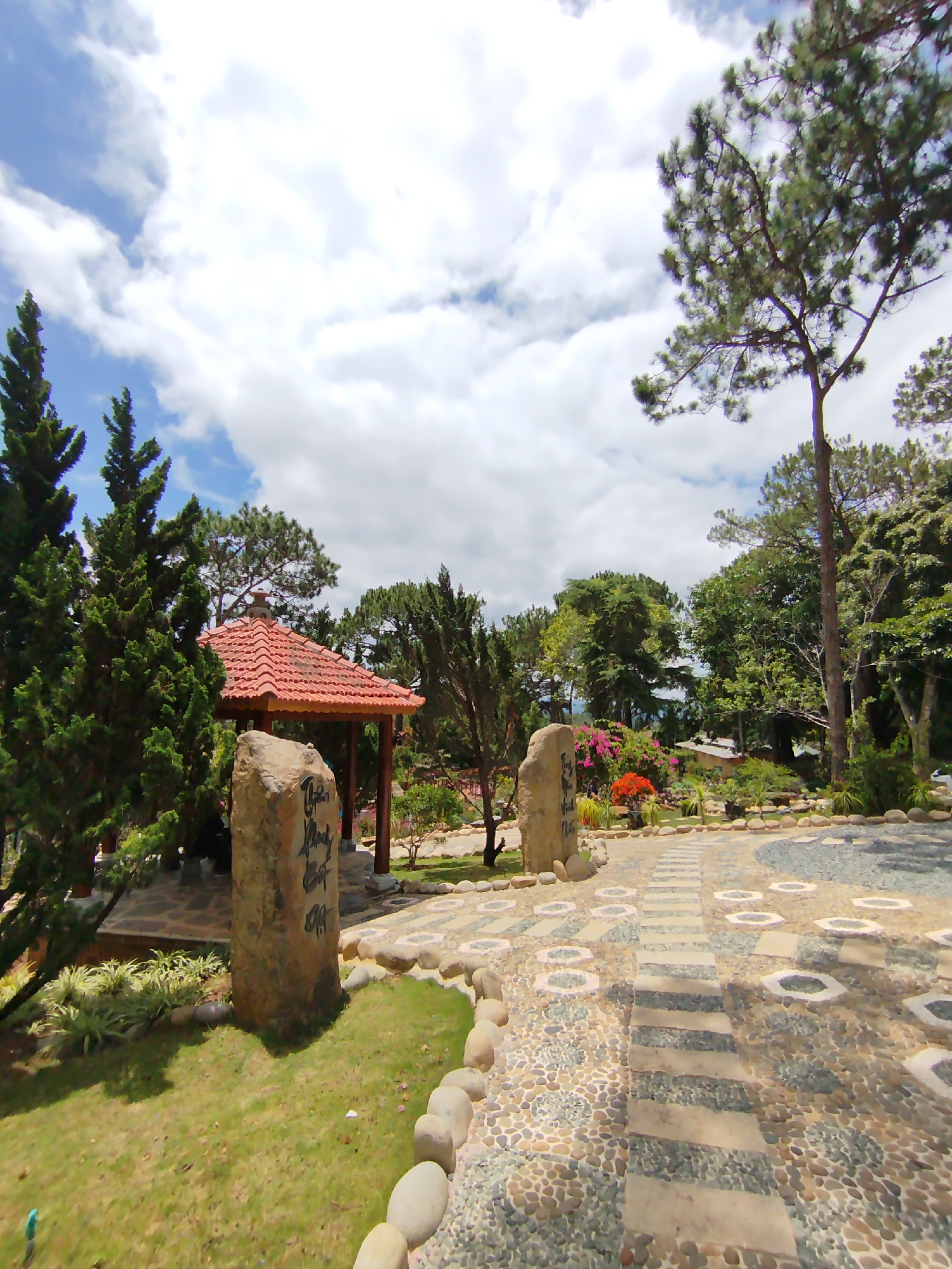 The image shows a garden scene, likely in Da Lat, Vietnam, characterized by its landscaping and architectural elements. A red-tiled gazebo is visible on the left, surrounded by lush greenery. In the foreground, there is a stone pathway lined with rounded stones, leading into the garden. Large, upright stones with inscriptions in Vietnamese stand at the sides of the path. The sky is partly cloudy. The vegetation includes a variety of trees and shrubs, and flowers can be seen in the background.