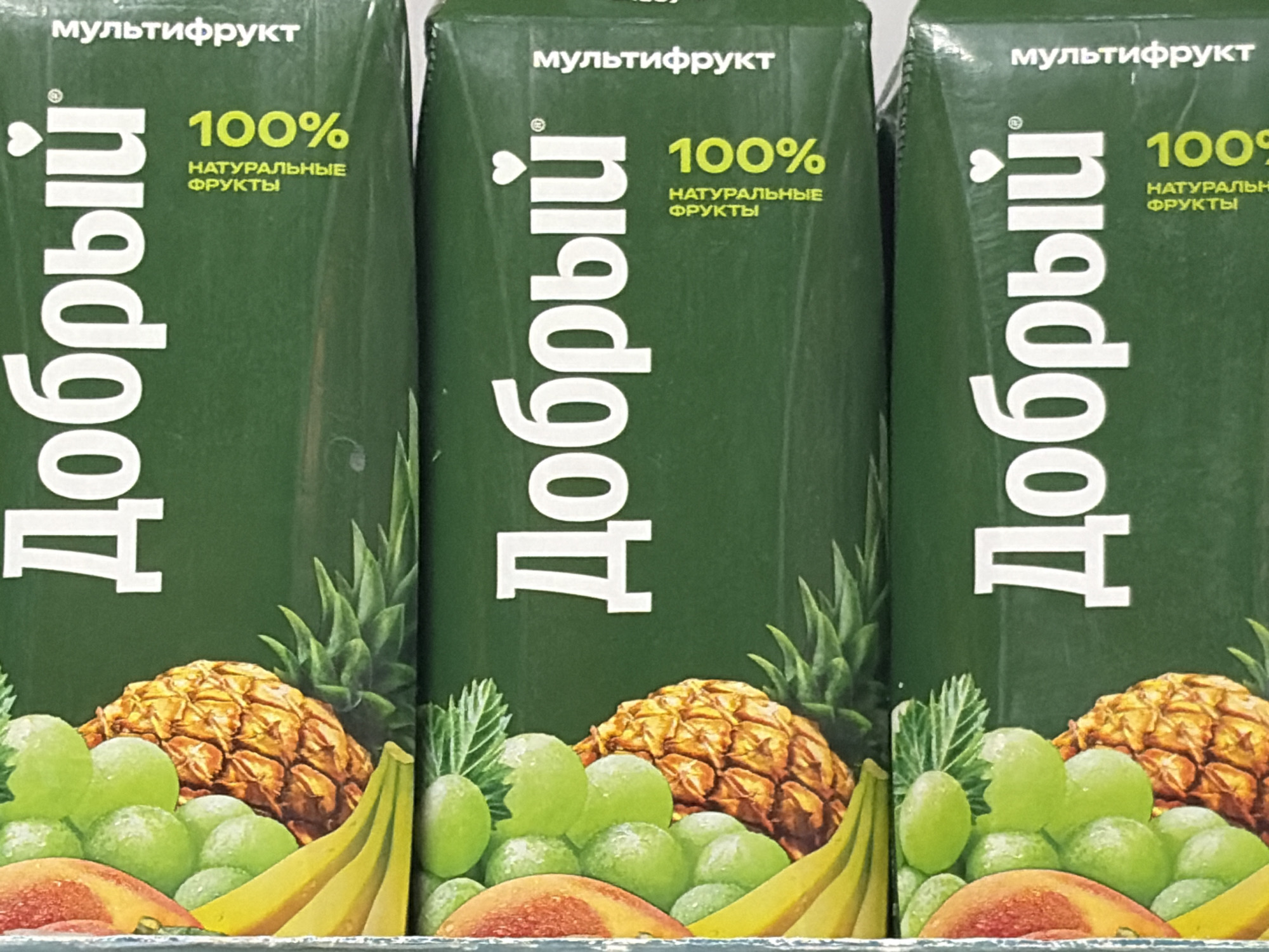 This media file features a close-up, eye-level shot of three identical juice cartons, stacked side-by-side. The cartons are primarily a deep green color, with prominent branding and imagery. Each carton displays the word "Добрый" vertically in large, white, sans-serif font, accompanied by a small red heart symbol above the first letter. Above this, the text "мультифрукт" is written horizontally. To the right of the vertical text, the number "100%" is displayed in bright yellow, with smaller text below it reading "НАТУРАЛЬНЫЕ ФРУКТЫ" (Natural Fruits) also in yellow.
The lower half of each carton is adorned with illustrations of various fruits, including pineapples, green grapes, and bananas. The realism of the fruit illustrations suggests a focus on natural ingredients.
The overall presentation is clean and emphasizes the product's natural qualities and 100% fruit content. The lighting appears even, indicating an indoor setting, possibly a retail environment like a supermarket shelf. No people are visible in the image, and the focus is solely on the product itself. The visual style is bright and appealing, designed to attract consumers. The visible text is in Russian. Given the location context provided, this is likely a product sold in Russia.