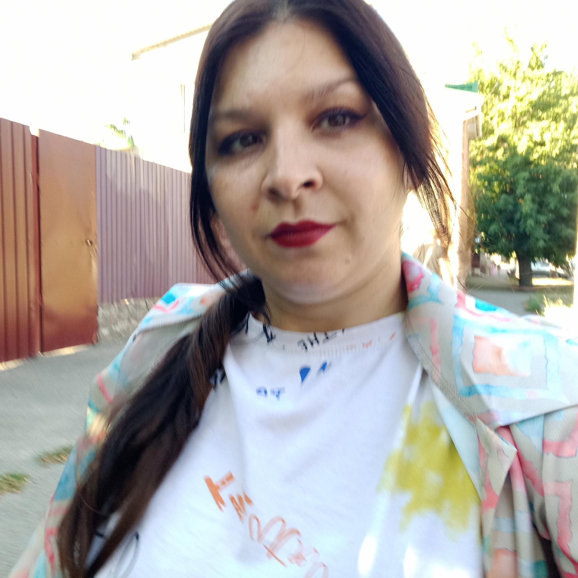 The image is a selfie taken outdoors, likely in an urban or suburban setting in Kropyvnytskyi, Ukraine, during daylight hours. The foreground is dominated by a woman with dark hair and makeup, including dark eyeliner and red lipstick. She is wearing a white t-shirt with some blue and orange lettering or drawings, and an open, light-colored jacket with a colorful, abstract pattern. Her hair is dark and styled in a braid that extends down her chest. Behind her, a textured, light purple fence runs horizontally. To the right, a large green tree is visible, and beyond that, a building with a green roof. The ground appears to be a paved surface, possibly a sidewalk or street, with some patches of dry grass. The lighting suggests it's a sunny day, casting some shadows and bright highlights. The overall scene depicts a casual outdoor moment.
