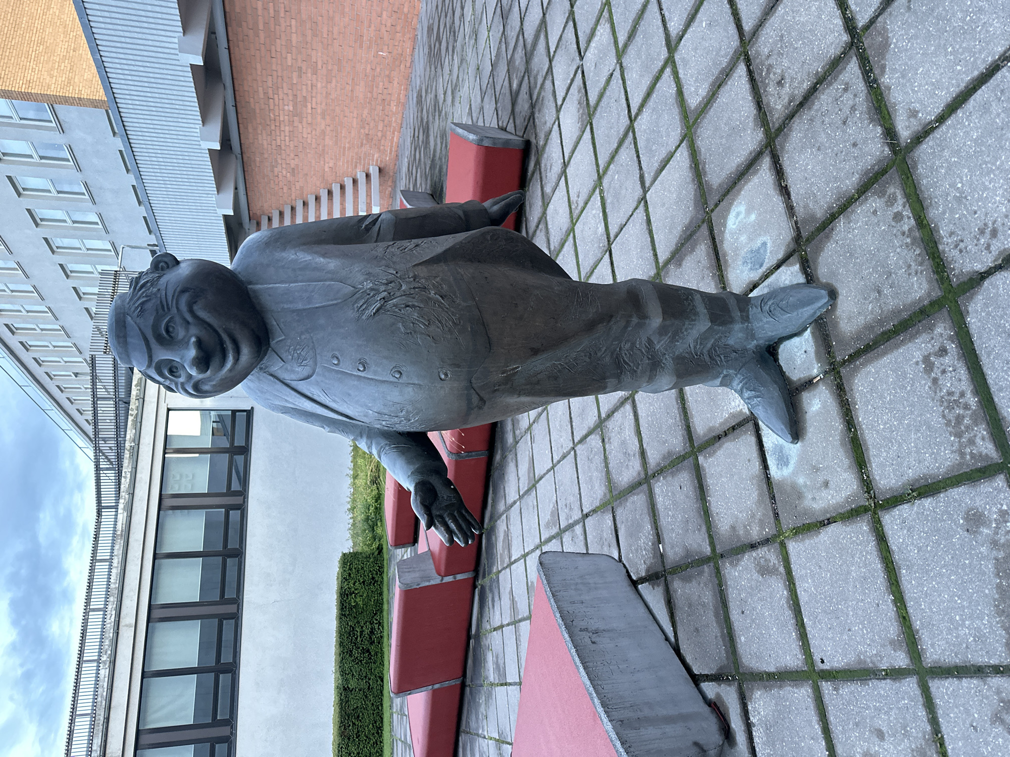 A bronze statue of a plump, grinning man in a suit stands on a square paved outdoor area. The statue is facing slightly to the left and has its right arm extended as if offering a handshake. Behind the statue, to the left, is a low, manicured hedge and a modern building with rows of windows. To the right of the statue, a brick building with an external staircase is visible. In the foreground and midground, several red, angular benches are placed on the paved area. The sky is overcast. The statue appears to be located in front of the Estonian Academy of Arts (EKA) in Tallinn, Estonia, a location known for its modern architecture and public art installations.
