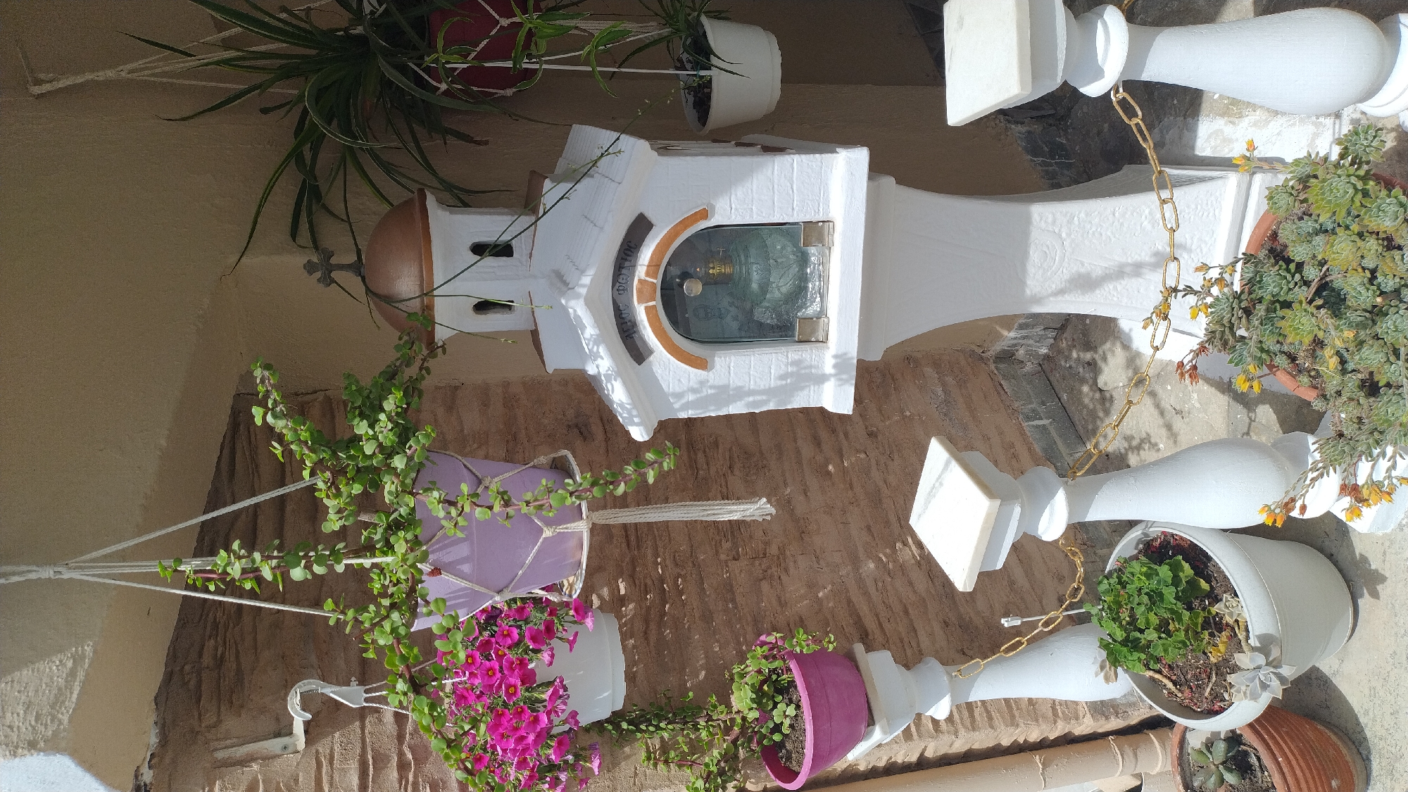 This image depicts an outdoor scene in Νέα Μηχανιώνα, Greece. The central focus is a small, white, church-like structure mounted on a pedestal. This structure has a domed roof with a cross on top and a glass-fronted opening displaying religious iconography, likely a saint or an icon, along with a glass oil lamp. Around the base of this structure, and on the ground, are various potted plants. These include succulents in terra cotta and white pots, and a flowering plant with pink blossoms hanging in a white pot suspended by macrame. Another hanging basket with a trailing green plant in a purple pot is also visible. The background features a textured, light brown brick wall and a pale, possibly stuccoed, overhang. White balusters connected by a gold chain are positioned in the foreground, suggesting a pathway or a decorative border. The lighting indicates it is daytime, with distinct shadows present. There are no people visible in the scene.