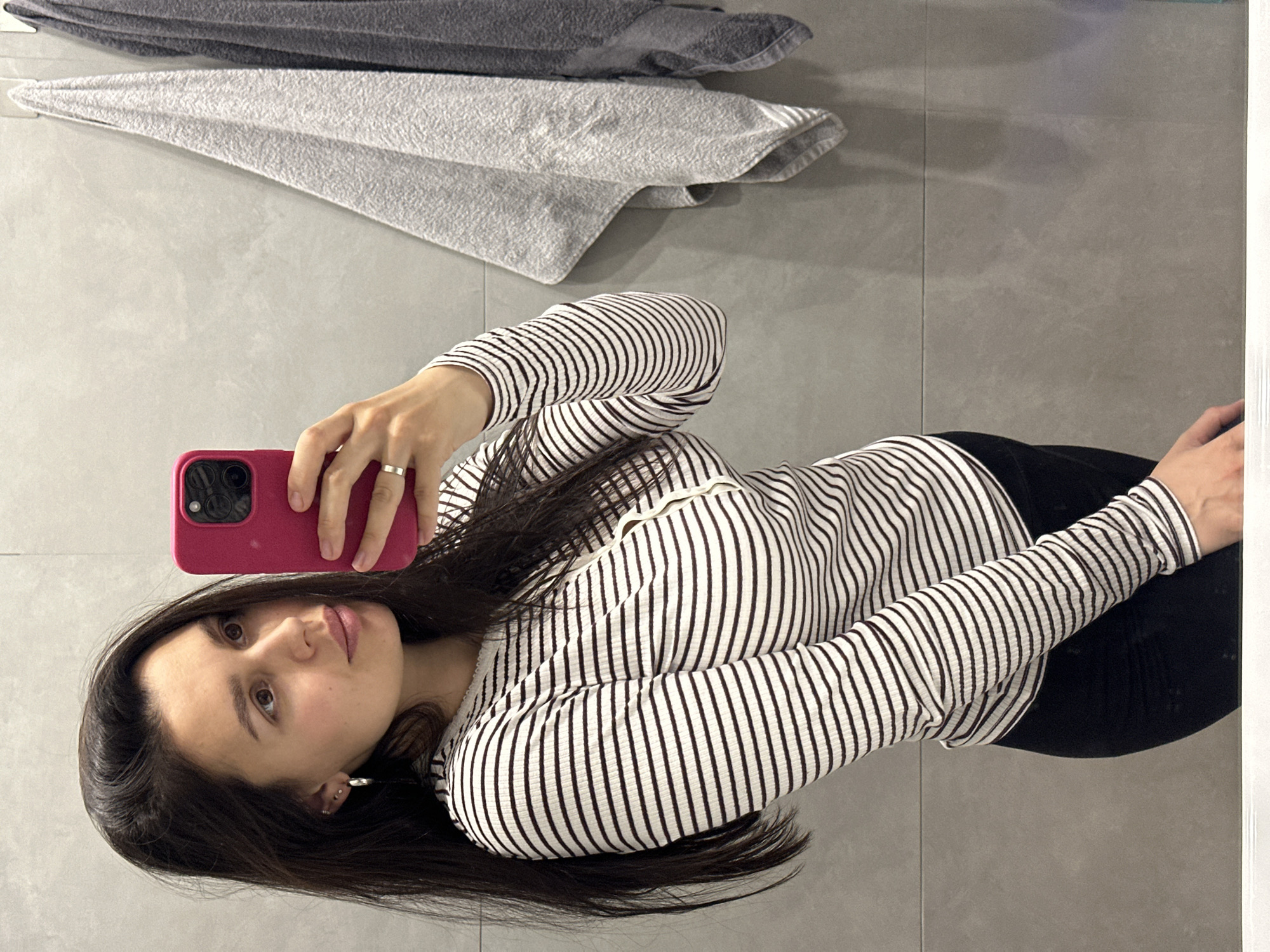 A woman is taking a selfie in a bathroom. She is wearing a striped long-sleeved shirt and black pants. She is holding a pink phone in her left hand. There are folded towels in the background. The bathroom has grey tiled walls. The location is not discernible.