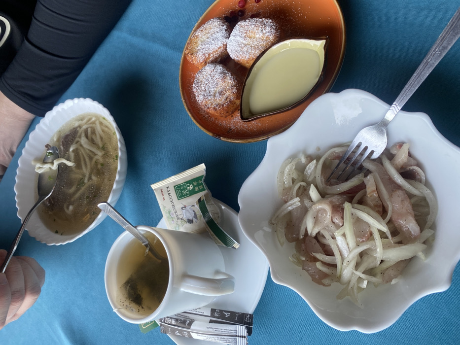 The image captures a meal consisting of soup with noodles in a white bowl, herring with sliced onions in another white bowl with a fork stuck in it, and small round cakes dusted with powdered sugar served with a dipping sauce on an orange plate. A cup of tea with a tea bag and sugar packets accompanies the food. The table is covered with a blue cloth. A person's arm is visible on the left side of the image.