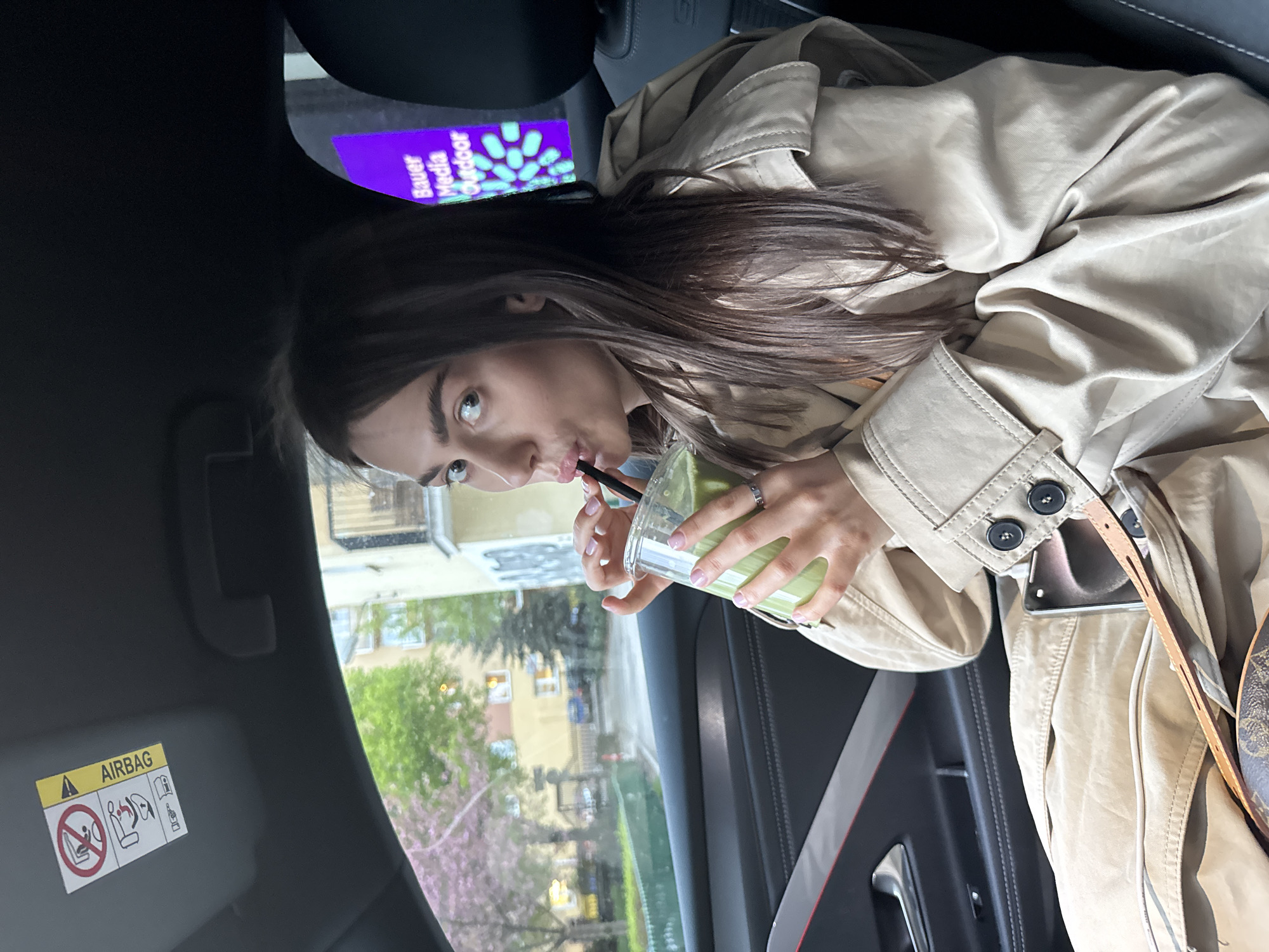 A young woman with light brown hair is seen inside a car. She is wearing a beige trench coat and drinking from a clear plastic cup with a black straw. The beverage in the cup is green. Her hands are holding the cup. A cell phone can be seen tucked into the pocket of her trench coat.
Outside the car window, a street view is visible with a yellow building, blossoming trees with pink flowers, and other greenery. Based on the "Bauer Media Outdoor" advertisement visible above her head and the surrounding architecture, this scene appears to be located in Warsaw, Poland. The car's interior is modern, with black trim and an "AIRBAG" label visible on the sun visor.