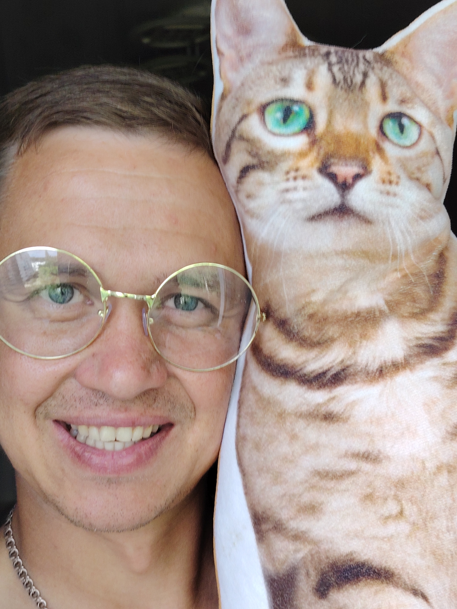 A man with short brown hair and glasses smiles and poses next to a pillow shaped like a cat's head. The man is wearing a silver chain. The pillow has green eyes and brown and tan fur. The background is dark. The image was taken in the city of Chaikovsky, Russia.