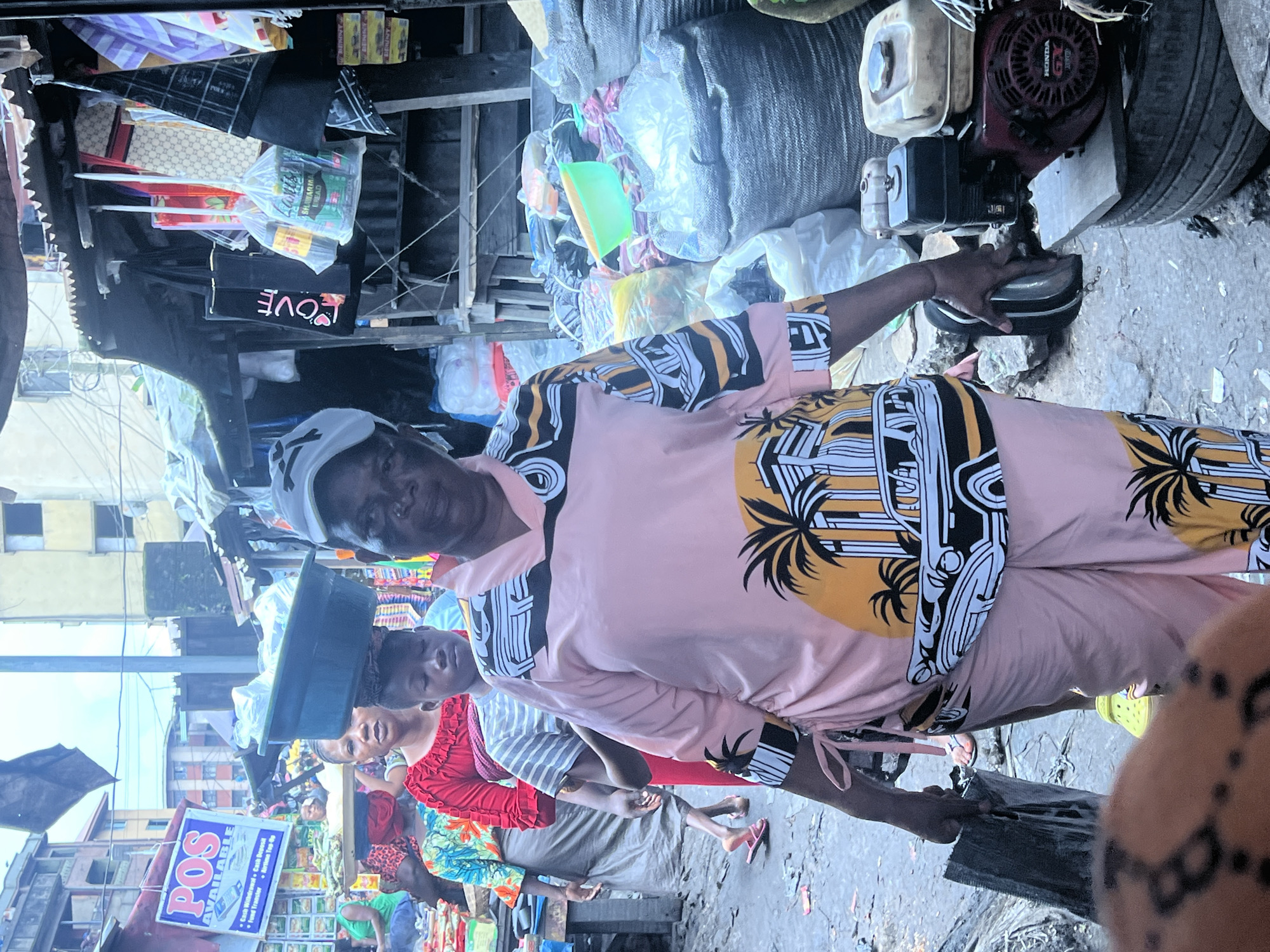 In Nkpor, Nigeria, a bustling market scene unfolds. A woman, her face obscured by a cap, walks towards the camera, her pink shirt adorned with a palm tree and retro car graphic. She carries a large, dark object in her right hand. Behind her, a young person with dark hair wears a striped shirt, and another woman in a red top with ruffled sleeves also moves through the market. Another woman, partially visible to the left, wears a colorful patterned shirt. The ground is dusty and uneven, with various items scattered around. Bags of goods, some in clear plastic, others in woven sacks, are piled high. A green plastic basin sits atop a larger basin on someone's head. A portable generator, branded "HONDA GX," is visible on the right side of the image, resting on stacked tires. The background reveals more market stalls and buildings, suggesting a dense urban environment. The overall impression is one of a lively, outdoor marketplace.
