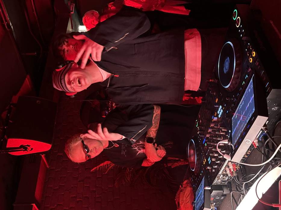 Two people pose next to a DJ setup under red lighting. The DJ has blond hair, sunglasses, and tattoos on their arm. The person next to them wears a striped hat and dark jacket. Both individuals have hands raised and are making gestures. A mixer and two turntables are prominent in the foreground, showcasing various buttons, screens, and cables. The background is dimly lit with a textured wall and hints of other people.