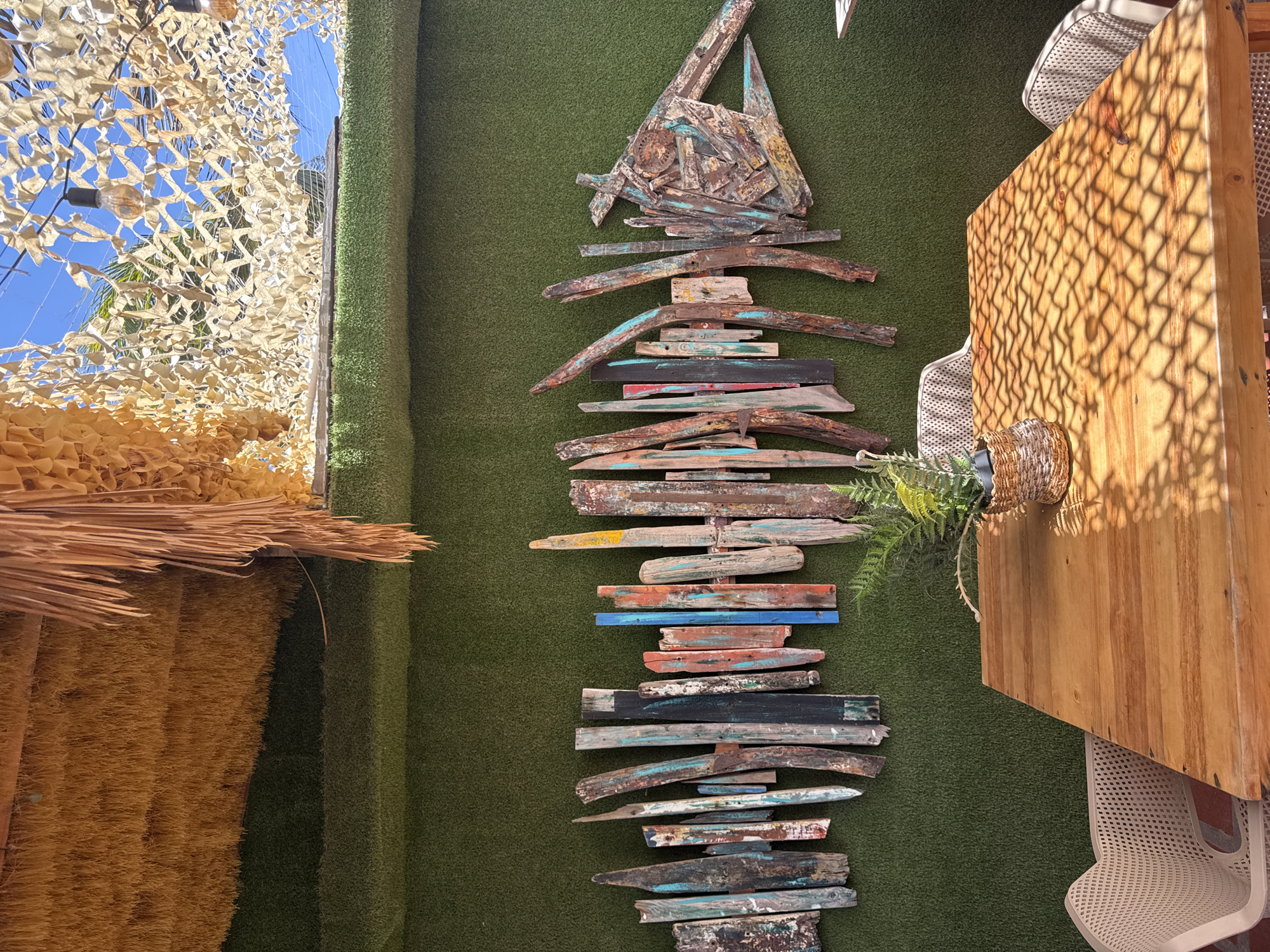 The image shows a wall decoration of a stylized fish made of driftwood mounted on a green artificial grass wall. The fish is constructed from various pieces of reclaimed wood in different colors and textures. Adjacent to the green wall is a structure with a thatched roof and a camouflage net extending above, suggesting an outdoor or semi-outdoor space. To the right is a wooden structure with shadows from a net-like material, likely a table or counter, with a chair partially visible beneath. The setting could be a restaurant or bar in Porlamar, Venezuela, given the coastal design elements. There are no people visible in the image.