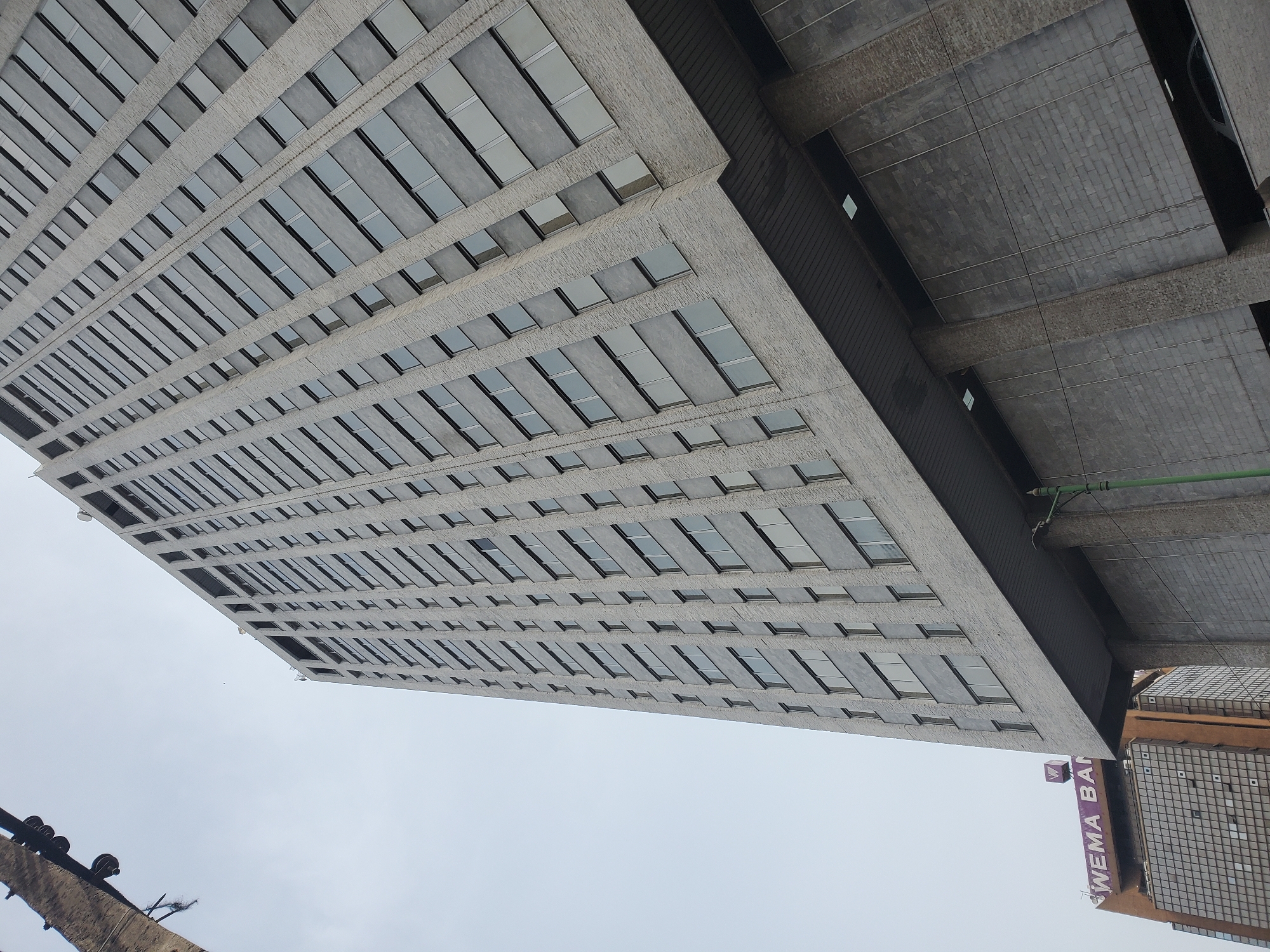 An upward-angled photograph captures an urban architectural scene, dominated by a tall, modern building on the right under an overcast sky. The primary structure is a high-rise building characterized by a light gray, textured facade, possibly aggregate concrete, with numerous rectangular windows arranged in horizontal bands. These windows appear somewhat reflective, showing subtle variations in light and shadow. The building extends vertically upwards, filling much of the right side of the frame.
To the left and upper-left, another structure, possibly an adjacent building or a lower section of the same complex, features a darker gray, heavily textured surface, resembling brickwork or concrete blocks. Visible beneath this section are large, dark structural beams and some small, square openings. A bright green pipe or conduit is attached to this darker structure.
In the bottom right corner, a portion of a distinct brown or orange building with a grid-like pattern of windows or facade elements is visible. Near this building, a white sign with purple lettering clearly displays "WEMA BANK" along with a purple "W" logo.
The sky, occupying the left side of the image, is a uniform light gray, indicating an overcast day with diffused lighting; no direct sunlight is apparent. In the far bottom-left, an indistinct, dark, angular object, possibly part of a crane or another construction element, is partially visible. The scene suggests a busy commercial or financial district within an urban environment, consistent with Lagos, Nigeria. No people or specific activities are depicted.