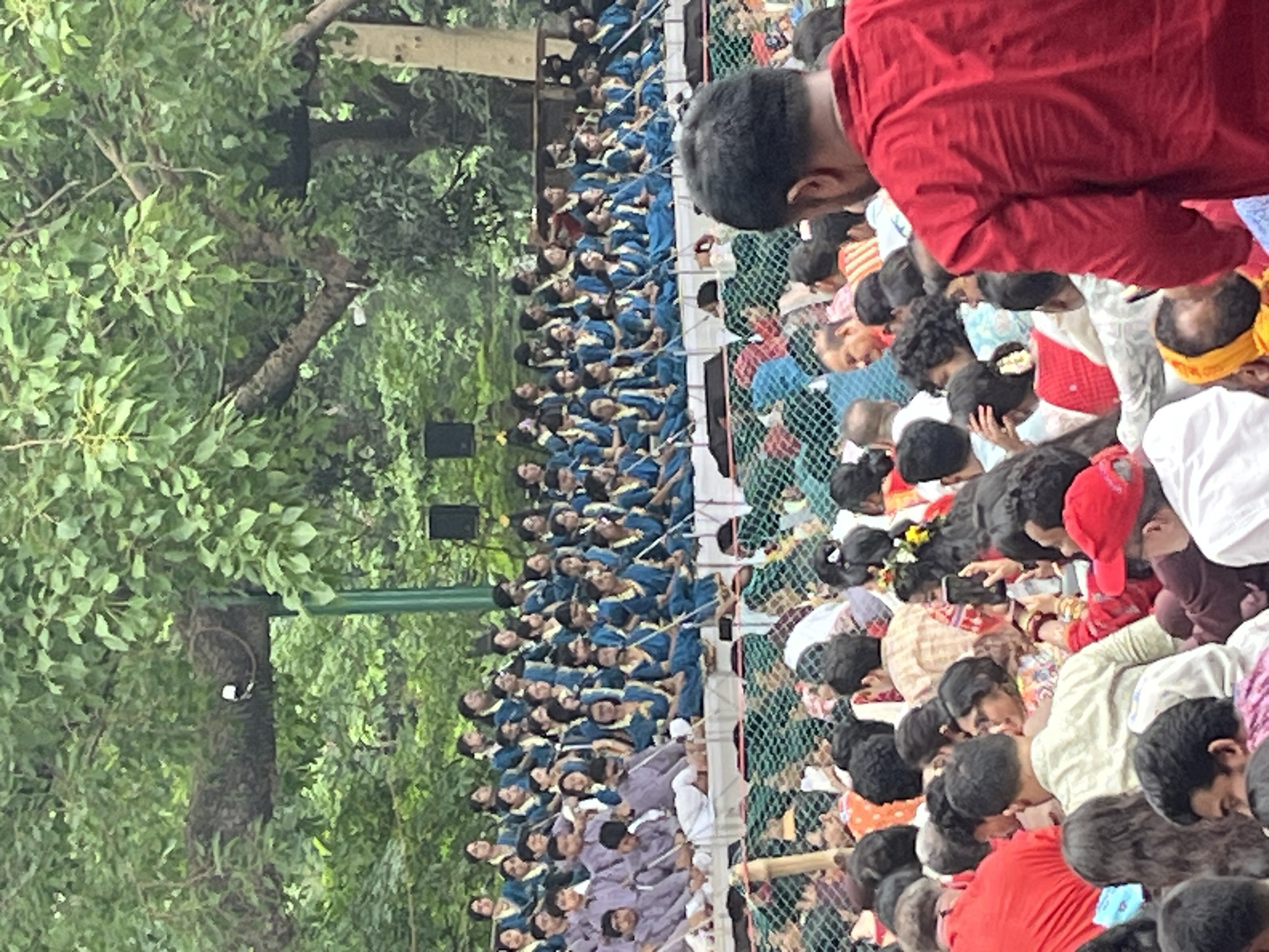The image shows a large crowd of people gathered outdoors, likely in Dhaka, Bangladesh. A line of individuals in blue uniforms sits behind a green chain-link fence. In front of the fence, a more diverse crowd is gathered, with some people in red shirts and others in various attire. Some individuals in the foreground are looking down, possibly at phones or other objects. A tree with dense green foliage is visible to the left of the crowd and fence. Speakers are mounted on a pole next to the seated people.
