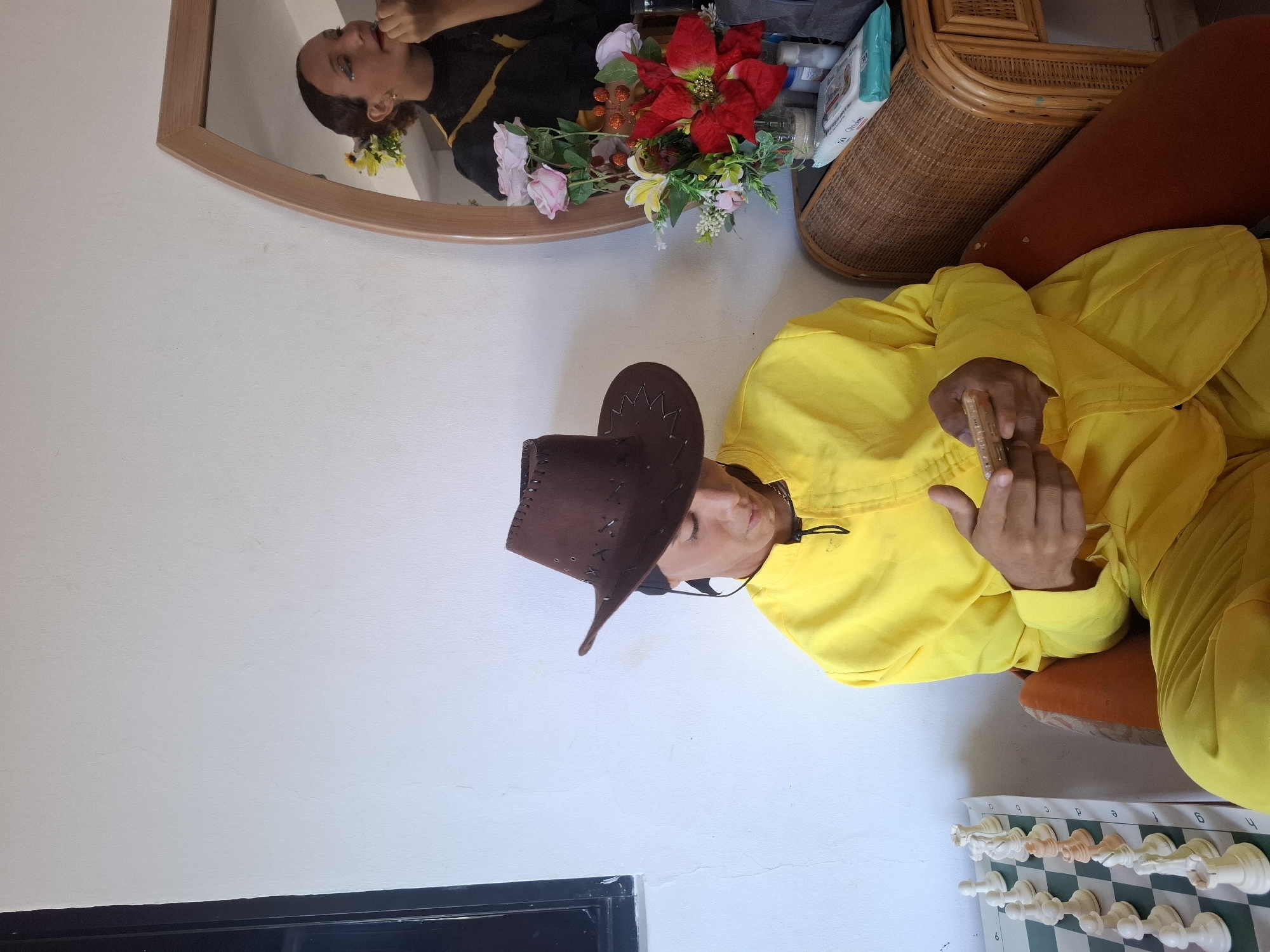 The image displays an indoor scene, likely a living room or common area in Playa El Agua, Venezuela, featuring two individuals and various household items. The main subject is a person seated on a rust-colored cushion or chair, adorned in a bright yellow long-sleeved tunic and a dark brown cowboy hat with decorative stitching. This individual is holding a smartphone with both hands and appears to be focused on its screen.
In the upper left, a large, curved wooden-framed mirror reflects another person's head and upper torso. This reflected individual, possibly female, is wearing a dark top with yellow accents and has small yellow flowers attached to their hair. They are seen looking intently at their reflection, seemingly applying makeup or adjusting their appearance.
In front of the mirror, a vibrant arrangement of artificial flowers is visible, including a prominent red poinsettia, pink roses, and other yellow and white blossoms. Behind this floral display, on a piece of wicker furniture, various bottles and a package of "50 Wipes" are stacked.
On the floor to the right, a chessboard with white and light orange chess pieces is set up, displaying column labels "a" through "h." The room features plain white walls and a dark baseboard along the bottom. The bright, even lighting suggests it is daytime. The scene conveys a relaxed, domestic atmosphere.