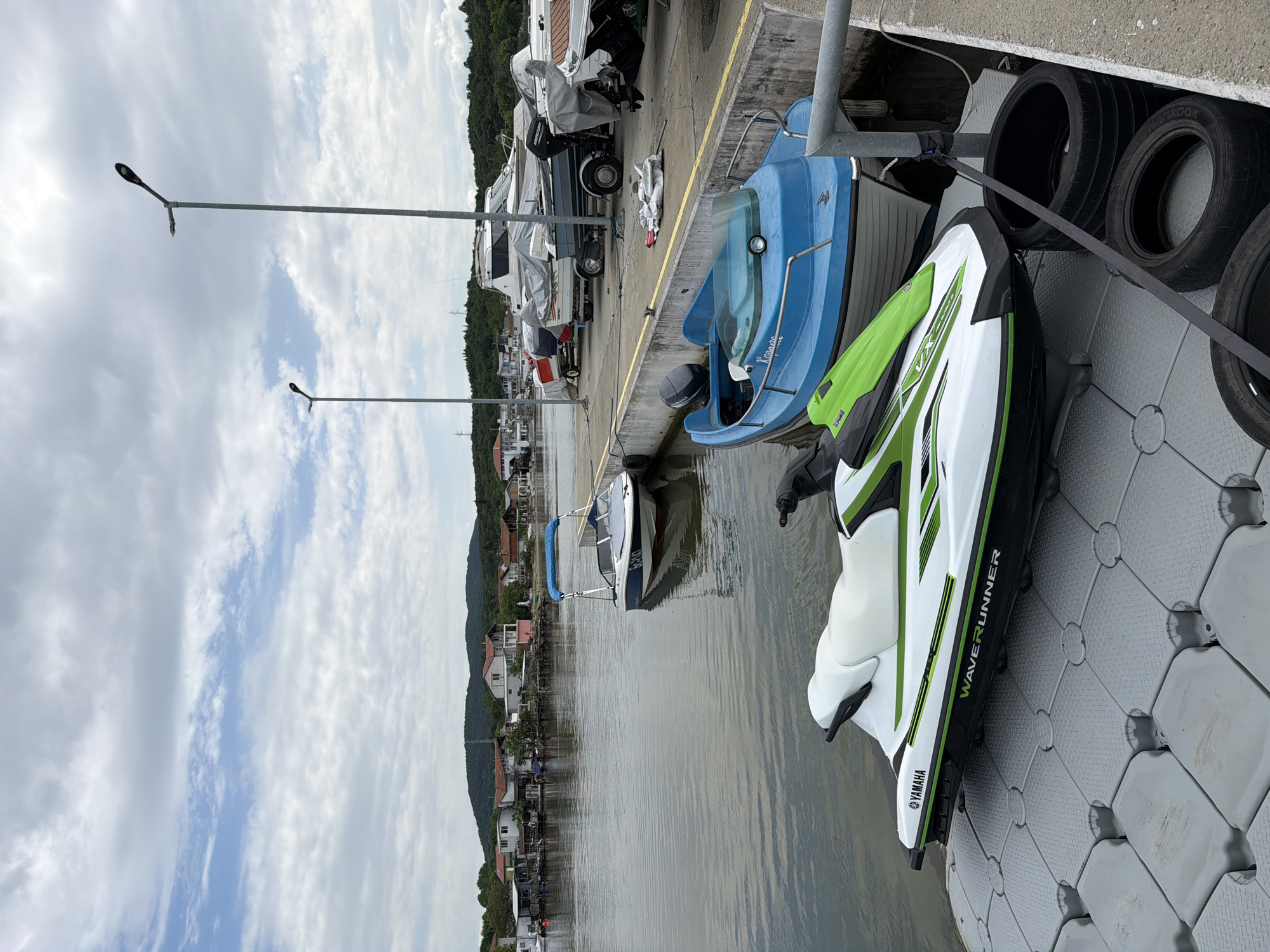 The image depicts a waterfront scene, likely a marina or dock, with several watercraft. In the foreground, a white and green Yamaha WaveRunner is secured on a gray floating dock, next to a small blue motorboat. Alongside the dock, several boats are moored, some covered with white tarps. The dock is lined with black tires used as bumpers. The background shows a coastal town with residential buildings and lush green hills. Streetlights are visible along the dock area. The sky is partly cloudy. The image shows the location in the city of Burgas, Bulgaria.