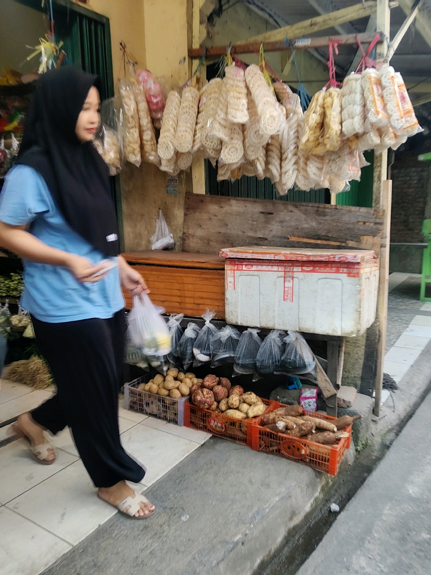 In a street-level view of a market stall in Mekarmukti, Indonesia, a woman wearing a black hijab and a light blue t-shirt walks from left to right, carrying a transparent plastic bag in her left hand and a white plastic bag in her right. She wears black pants and tan slip-on sandals. The stall features an assortment of goods. Hanging from a wooden structure above are numerous bundles of packaged snacks, primarily round crackers and fried snacks in various shades of white and yellow. Below the snacks, a weathered wooden counter displays a white cooler box with red trim and "FRAGILE" labels, situated next to a wooden drawer. In front of the counter, several grey plastic crates hold potatoes and other root vegetables. A concrete sidewalk runs along the right side of the image, with a gutter. The background shows a building facade and a green metal structure.