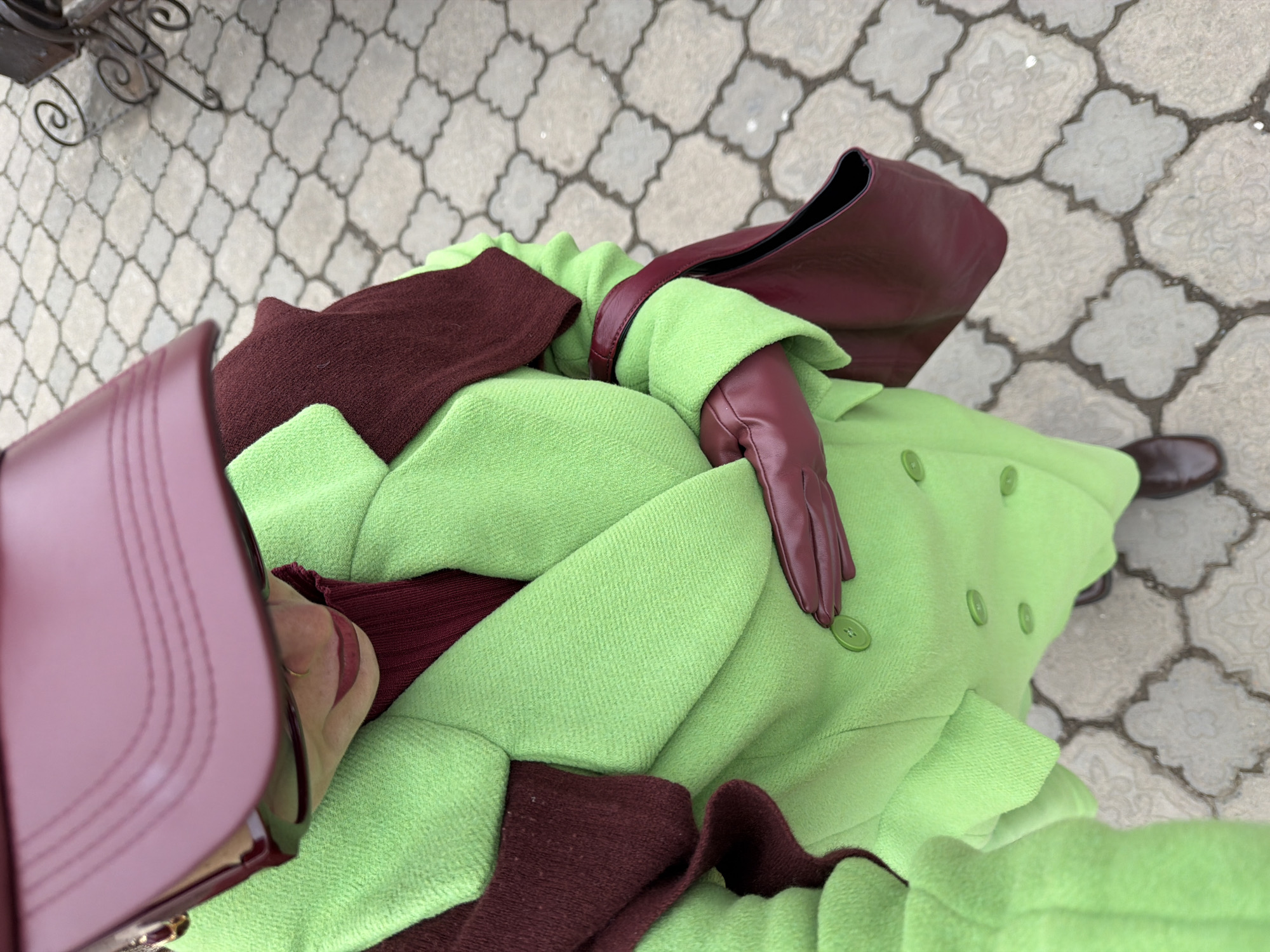 The image, taken from an overhead perspective, shows a person wearing a bright green wool coat with matching buttons. The coat features a burgundy-colored wool trim around the collar and lapels. The person also wears a pair of burgundy-colored leather gloves. A burgundy leather handbag is held near the person's head.
The person stands on a paved area featuring interlocking gray stone tiles. The cobblestone ground extends into the background. The overall scene suggests an outdoor location in an urban environment.
Based on the style of clothing and location of the image being sourced from Krasnoyarsk, Russia, it is possible the scene is a public area in Krasnoyarsk, Russia, although no clear landmarks or visual cues exist in the image to positively identify the specific spot.