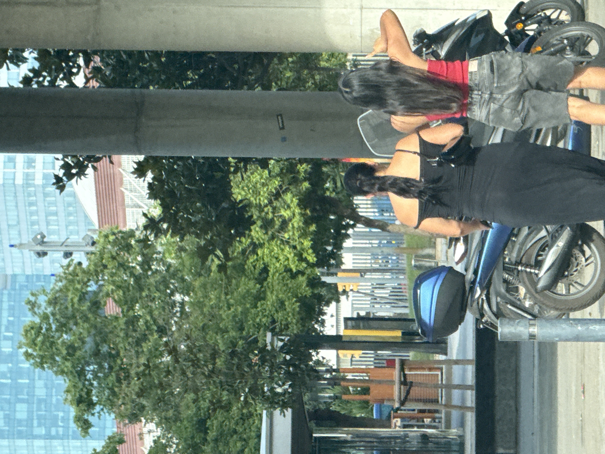 Two women are standing next to a motorcycle in an urban environment, with buildings and trees visible in the background. The woman on the left is wearing a black off-the-shoulder dress and holding a black purse. The woman on the right is wearing a red tank top and denim shorts. The motorcycle is blue and black. The scene is lit by daylight. The location is indicated to be in L'Hospitalet de Llobregat, Spain.
