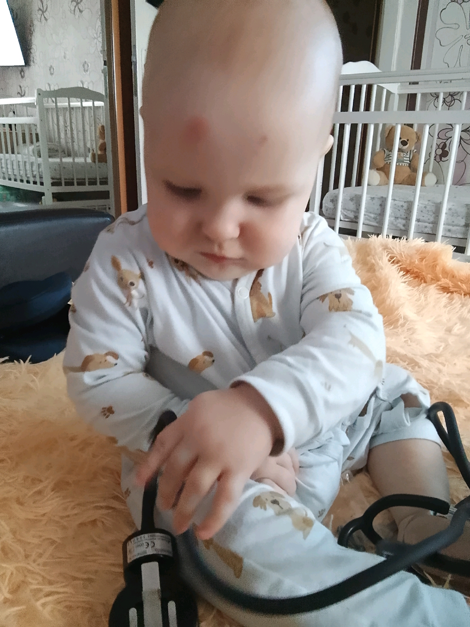 An infant with a small red mark on its forehead is sitting on a fluffy orange surface in front of a white crib. The infant is wearing pajamas with a pattern of small animals. A stethoscope is visible in front of the infant, with one of its hands near it. In the background, a white crib with a teddy bear is visible. The scene appears to be indoors, possibly a bedroom, and based on the provided context, is located in Minsk, Belarus.