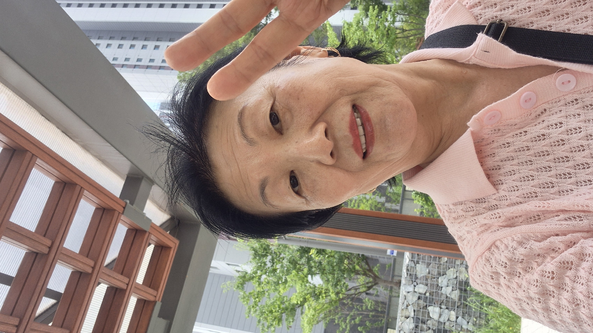 A medium shot selfie captures an elderly East Asian woman with short black hair, fair skin, and smiling lips, raising her right hand in a peace sign. She is wearing a pink knit polo shirt. The background includes a modern building and lush green trees, suggesting an urban setting. The overhead structure with wooden beams and translucent panels, along with the building in the background, are indicative of an outdoor public space in Ansan, South Korea.