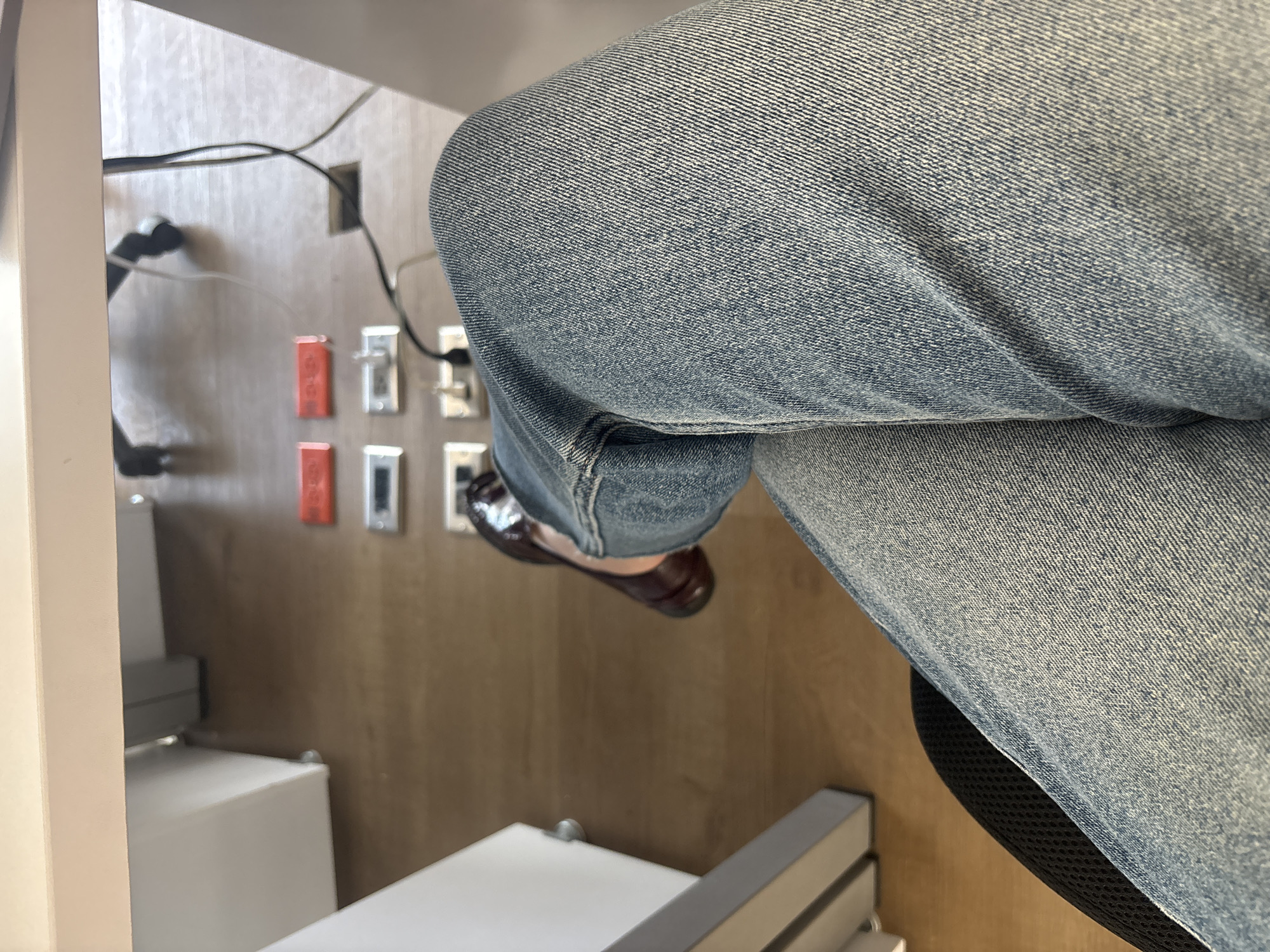The image shows a person wearing blue jeans sitting with their legs crossed. A brown shoe is visible on the lower leg. The setting appears to be indoors, with a light-colored wooden wall featuring electrical outlets and cables. Parts of a white structure, possibly a desk or cabinet, are also visible. There's no information available in the image to suggest it was taken in the city of Buenavista, Mexico, or to identify any actions, interactions, or subjects beyond what is visually present.
