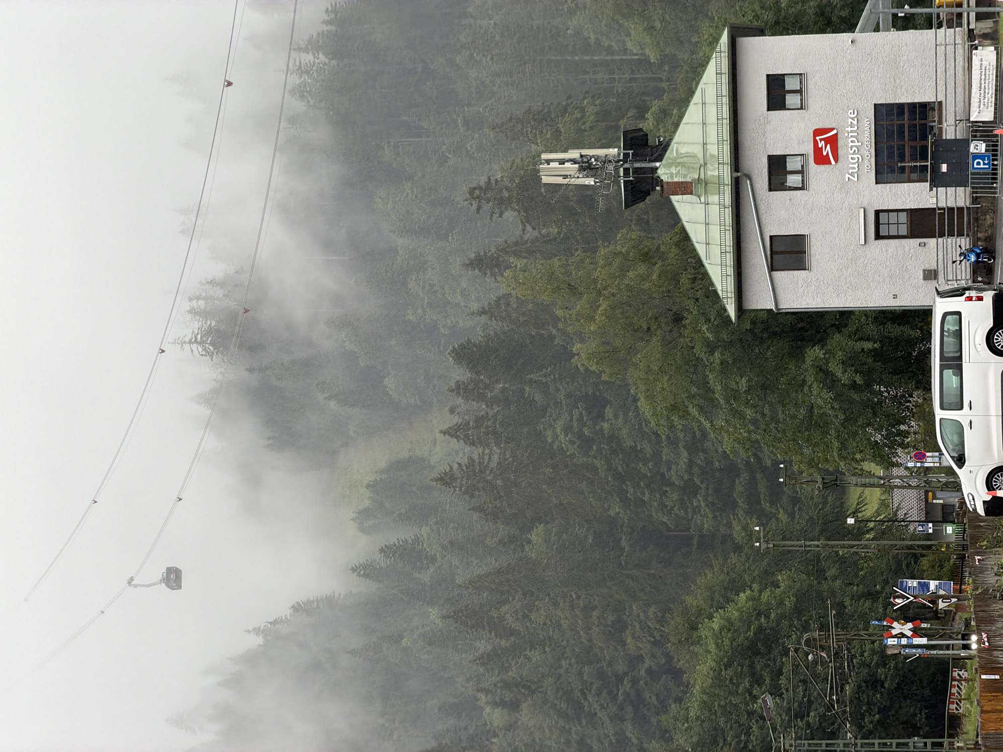 The image shows a cable car station building in Grainau, Germany, likely at the base of the Zugspitze cable car line. The building is a light-colored, two-story structure with a green, sloped roof. Above the building, a prominent logo with the text "Zugspitze TOP OF GERMANY" is visible. The building also features several windows and a dark-colored door. A white minivan is parked in front of the building, and a small blue scooter is parked to the right of the entrance. To the left of the building, there are train tracks, signal poles, and various road and railway signs. In the background, a lush, dense forest covers a steep mountain slope, partially obscured by fog. A cable car with a cabin is suspended by thick cables, traversing the foggy mountain landscape. The sky is overcast, and the overall atmosphere is muted and hazy, suggesting a cloudy or foggy day.
