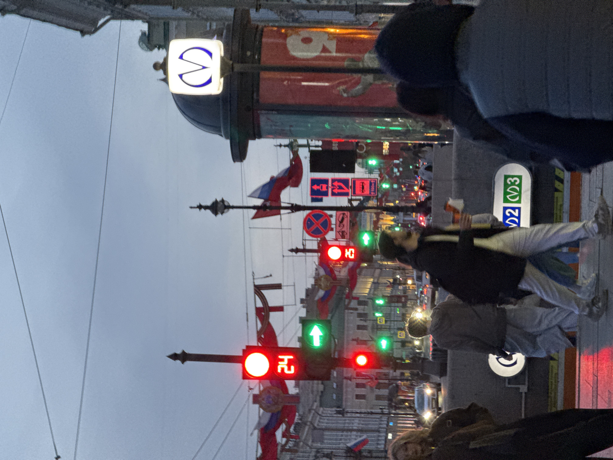 The image captures a street scene in Saint Petersburg, Russia, at twilight. The sky is overcast. The primary subjects are people walking on the sidewalk. A pedestrian in a dark jacket and light pants is in motion. A woman is in the foreground. Traffic lights are visible, displaying red lights in the horizontal axis, and a green light is indicated in the vertical axis. Russian flags are displayed from the poles. Prominently, the Saint Petersburg Metro symbol (a stylized "M") is illuminated on the station entrance. A sign with route numbers, “2” and “3” are shown near the ground. The building in the background is adorned with numerous small lights. A variety of traffic signs are posted on the poles. Overhead wires are visible against the sky.