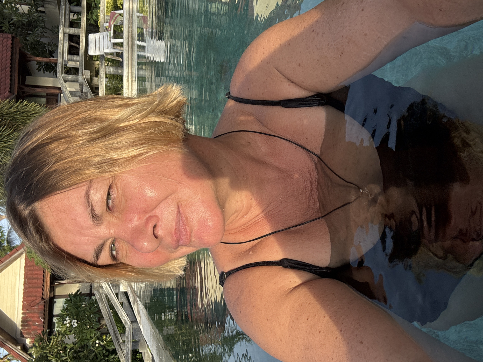 A woman with blonde, shoulder-length hair is in a swimming pool. She is wearing a black swimsuit and a necklace. The water reflects the sunlight. In the background, there is a building with a red roof and a white chair. The setting appears to be a resort in Ban Bang Rak, Thailand.