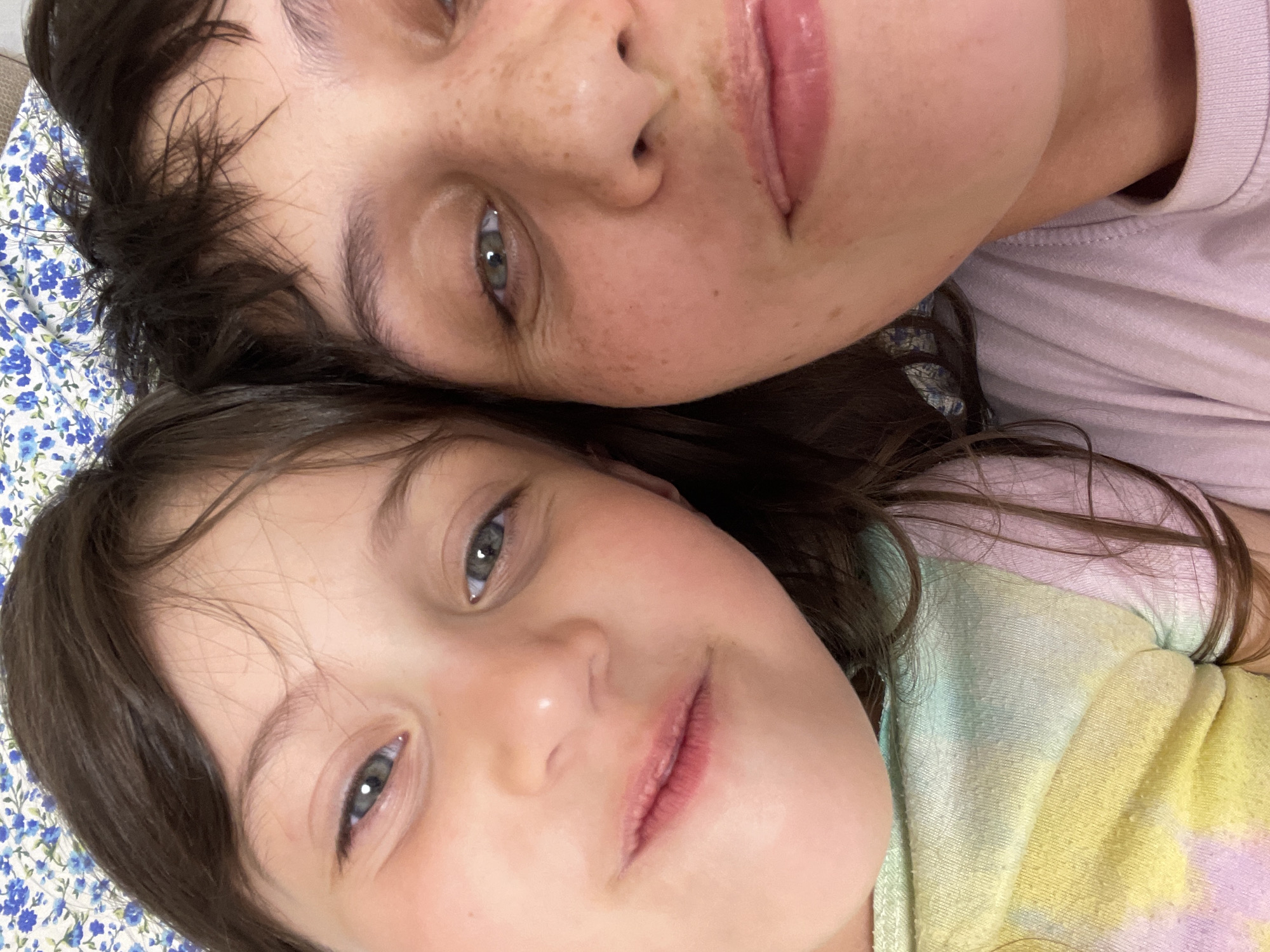 Two individuals, a child and an adult, are closely positioned together, likely lying down. The child has brown hair and light-colored eyes. The adult has dark hair, fair skin with freckles, and light-colored eyes. The child is wearing a tie-dye colored shirt, while the adult is wearing a light purple shirt. The background includes a floral patterned fabric. The location is not discernible from the image, and there is no indication the scene is in Izhevsk, Russia.
