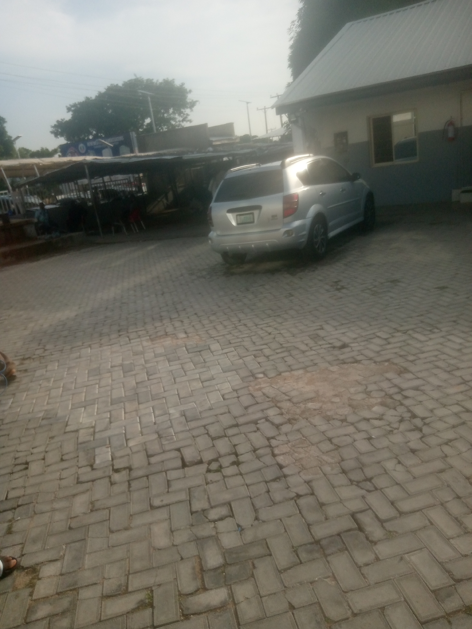 This media file depicts an outdoor scene in Jalingo, Nigeria. The foreground is dominated by a paved area made of interlocking grey bricks, some of which appear worn or discolored. To the right, a silver SUV is parked, facing away from the viewer, with its rear bumper and taillights visible. The car is parked next to a building with a light-colored facade, a darker grey band running horizontally across it, and a white metal roof. A window with a dark frame is visible on the building.
Behind the SUV and the building, a covered outdoor structure with a dark roof supported by poles is visible. Underneath this structure, several dark shapes, possibly furniture or stalls, are present. In the background, a large green tree with a sprawling canopy stands prominently. A blue sign with white text is partially visible behind the tree, and a telephone pole with wires is also present. The sky is overcast, suggesting a cloudy or hazy day. The lighting indicates it is likely daytime, possibly late afternoon or early morning due to the soft light. A person's foot wearing a sandal is visible in the lower left corner of the frame. The overall impression is a scene in a public or commercial area, perhaps a marketplace or an establishment with parking facilities.