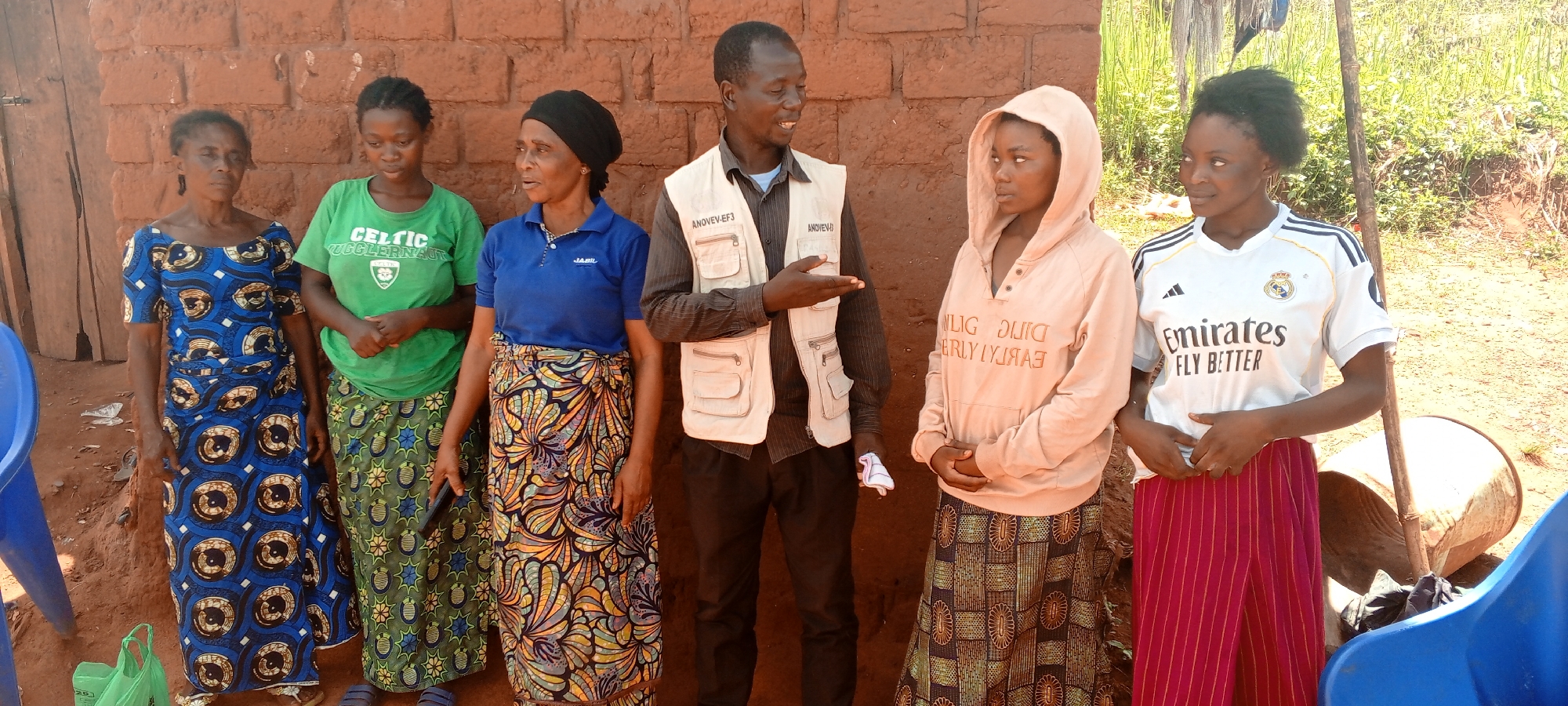 This outdoor scene in Makungu, Democratic Republic of the Congo, features six individuals: five women and one man, gathered against a backdrop of a reddish-brown mud or brick wall. The lighting suggests it is daytime with clear weather.
The man stands slightly right of center, wearing a light-colored, multi-pocketed vest over a dark shirt and trousers. He is actively speaking, gesturing with his right hand towards the women on his left. Visible text on his vest includes "ANDREW" on one pocket and "ANOVEV-EF3" on others.
To his immediate left, a woman in a black head covering, a blue polo shirt with "JABIL" printed on it, and a colorful patterned skirt appears attentive, looking towards the man. Next to her, a woman in a green "CELTIC JUGGERNAUT" t-shirt and a green patterned skirt holds a black mobile phone. On the far left, an older woman wears a blue patterned dress.
To the man's right, two younger women stand. One wears a light peach-colored hooded sweatshirt with reversed text (appearing as "NULIG OUIG ERLYIYJRAE", possibly a mirrored phrase like "EARLY YEARLY GIVING") and a dark patterned skirt, looking away. The other, on the far right, wears a white Real Madrid jersey with "adidas" branding, the club crest, and "Emirates FLY BETTER" text, paired with red striped trousers, also looking away.
The ground is reddish-brown soil. A blue plastic chair is partially visible on the far left, and another on the far right, near a light-colored barrel lying on its side. A green plastic bag is on the ground near the first woman. The overall impression is an informal meeting or discussion within a rural community setting.
