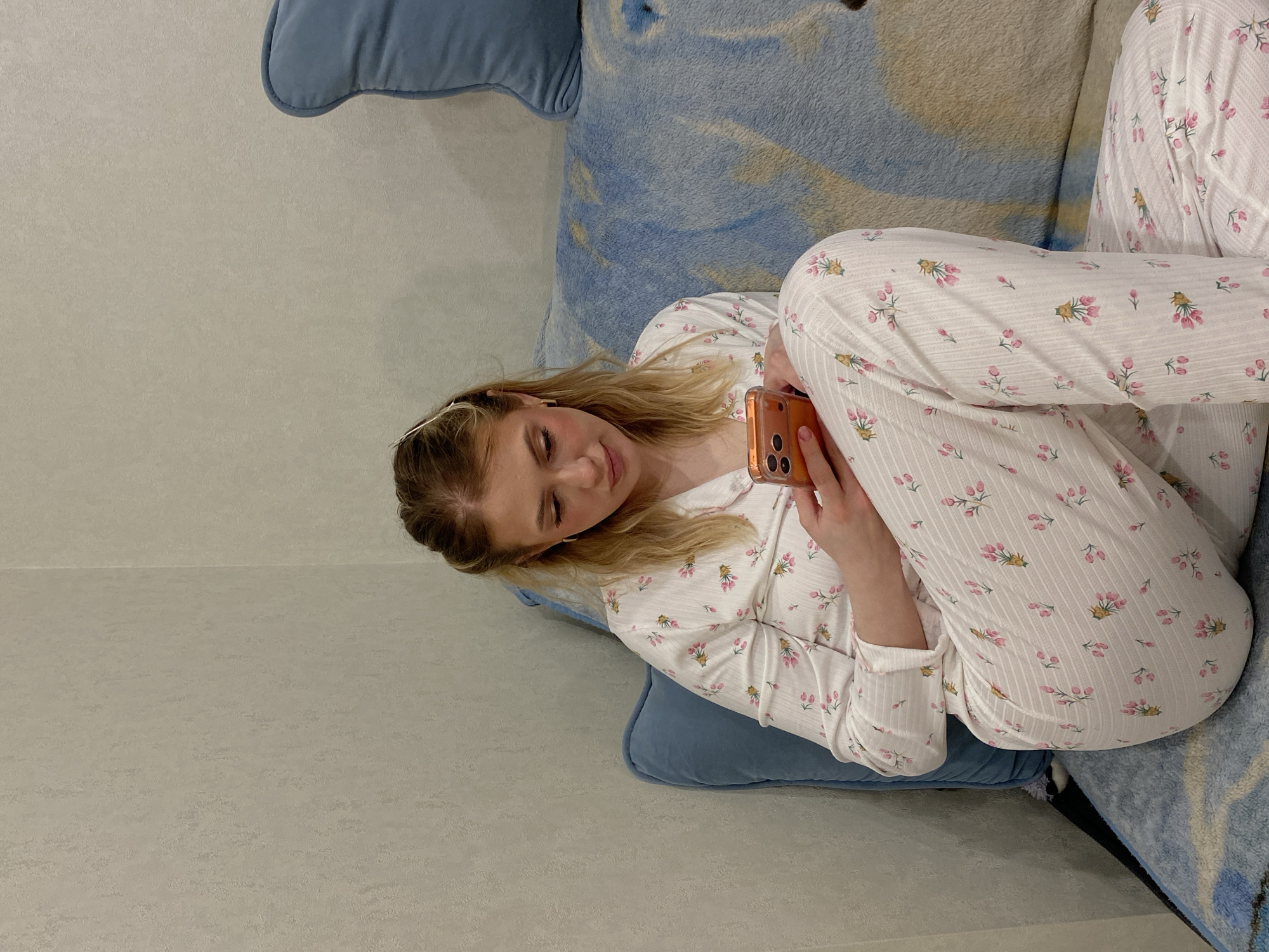 A young woman wearing floral pajamas is lying on a blue couch and looking at an orange smartphone. The location appears to be indoors, with a light gray wall visible in the background. The city of Kazan, Russia, cannot be determined from this image.