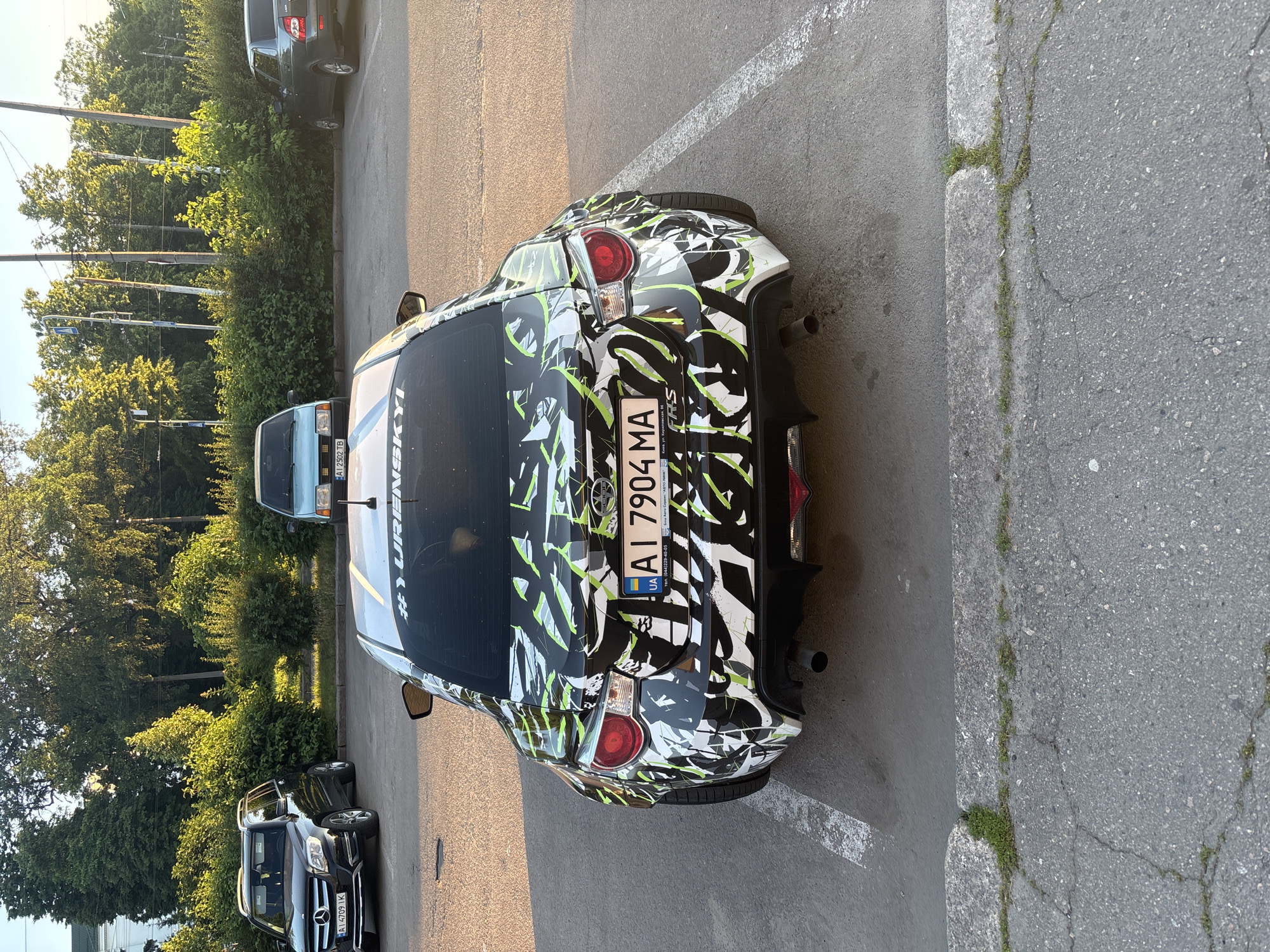 A black Subaru BRZ with a white, black, and green graffiti-style wrap is parked in a lot. The car has a custom license plate that reads "AI 7904 MA". A hashtag "#YURENTSKYI" is visible on the rear window. Other cars are parked nearby, including a black Mercedes-Benz SUV and a light blue sedan. The scene appears to be a parking lot in Bila Tserkva, Ukraine, with trees and overhead wires visible in the background. The lighting suggests it is late afternoon or early evening, with sunlight casting long shadows.