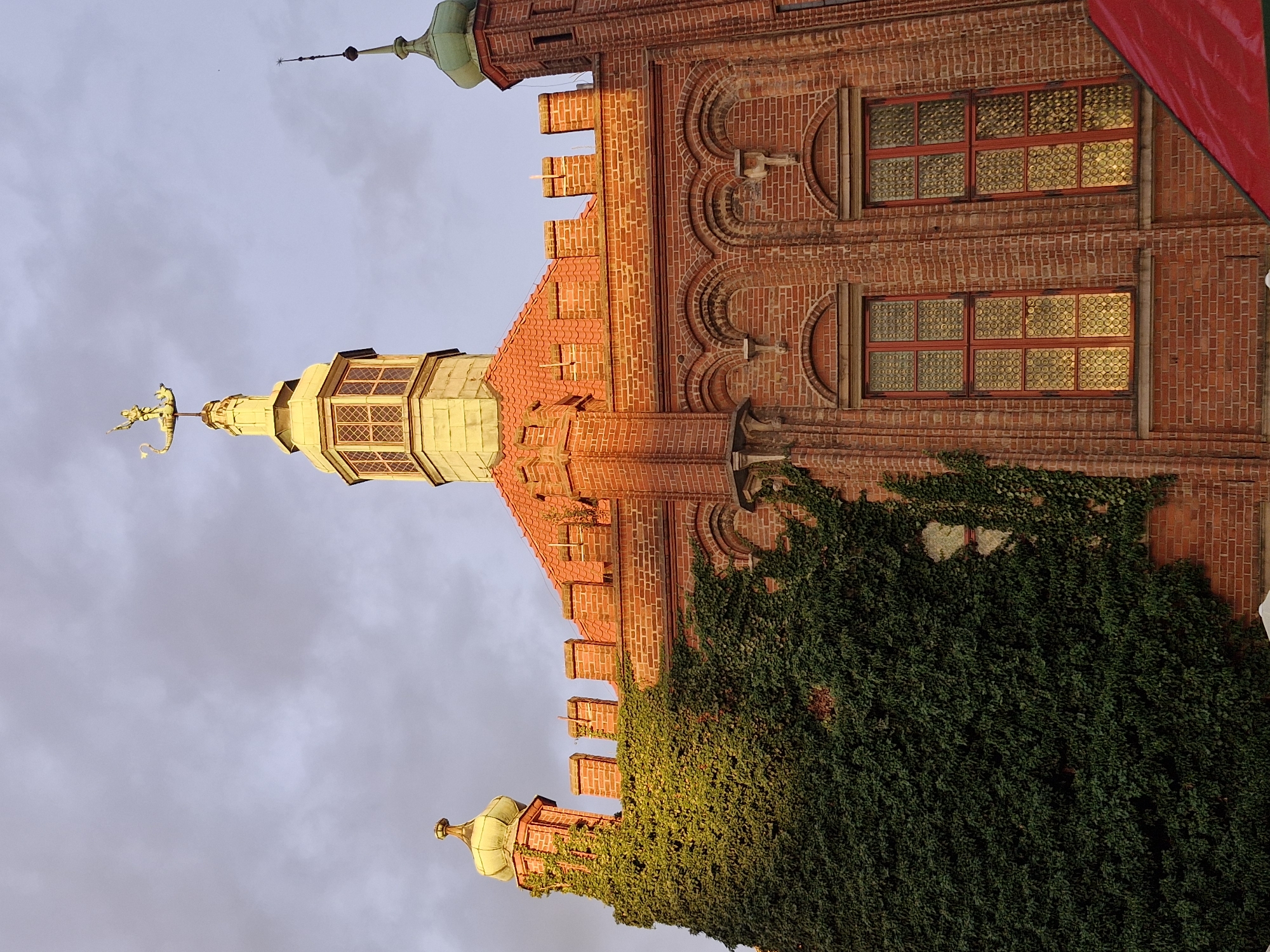 The image shows a close-up, low-angle view of the facade of the Great Mill in Gdańsk, Poland. The building is constructed of red brick and features a prominent tower with a golden weather vane depicting a ship and a figure. The tower is topped with a hexagonal lantern. To the left of the main tower, a smaller turret with a green dome is visible, partially covered by dense green ivy. The building has a red-tiled roof and crenellated battlements. Two tall, arched windows with red frames and distinctive patterned glass are visible on the right side of the building. The sky is overcast with soft, diffused light, suggesting it may be late afternoon or early morning. A portion of a red tent or awning is visible in the lower right corner of the image.