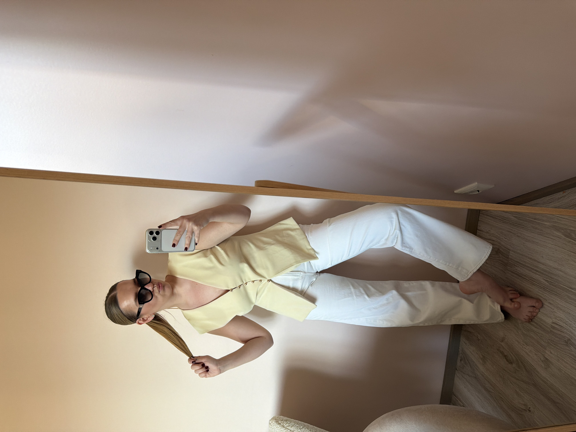 A woman is reflected in a mirror, taking a selfie with her smartphone. She is wearing sunglasses, a pale yellow sleeveless top with button details, and white trousers. She is pulling at her blonde ponytail with her left hand. The background consists of light pink walls and a wooden floor. The location is within a room, details of which are insufficient to establish with certainty if it is located in Dreiliņi, Latvia.
