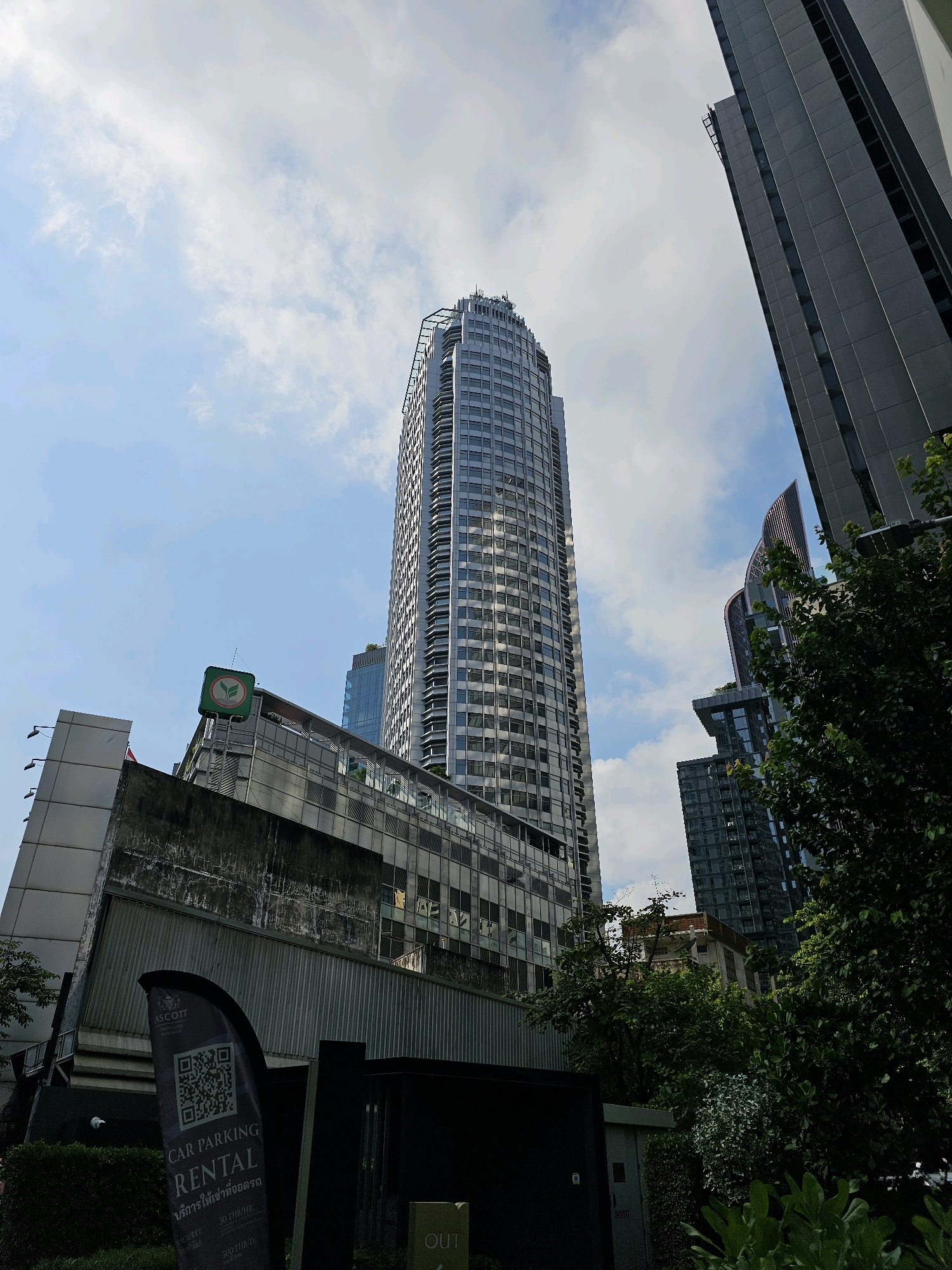 This is a low-angle, wide shot of a modern cityscape in Bangkok, Thailand, under a partly cloudy sky. The primary focus is a tall, slender, curved skyscraper with a reflective glass facade, appearing to be a residential or office building. To its right, another imposing, rectangular skyscraper with a gray facade dominates the upper right portion of the frame.
In the foreground, a weathered, concrete structure with visible signs of age runs horizontally across the lower half of the image. On top of this structure, a green square sign with a stylized leaf logo and Thai script is visible. Below this, a black, curved flag stands upright, displaying a QR code and text that reads "ASCOTT CAR PARKING RENTAL" in English and Thai. The flag also shows prices in Thai Baht. Lush green trees and foliage are visible on the right side of the frame, partially obscuring some of the buildings and adding a natural element to the urban environment. The overall scene suggests a busy, developed area within Bangkok, characterized by a mix of modern high-rise architecture and greenery. There are no people visible in the image.
