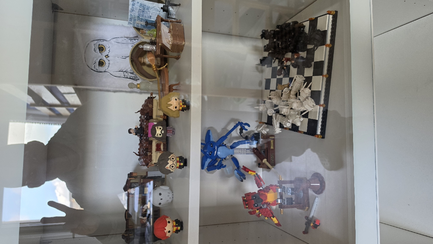 This image captures a brightly lit display cabinet showcasing a collection of brick-built models and figurines, likely taken indoors in Ihringshausen, Germany, during the daytime.
The left section of the cabinet is dedicated to Harry Potter-themed items. On the back panel, a drawing of Hedwig, the white owl, is visible. Arranged below are several Harry Potter characters in a Funko Pop-like or Lego mini-figure style, including Harry, Hermione, and Ron, some seated at a small table. Accompanying these figures are miniature accessories such as a broomstick and potion bottles. A small newspaper clipping displaying the text "DAILY PROPHET" is also part of the setup. Further down on this shelf, additional figures are displayed, including a white, moon-shaped character and a red-haired character.
The right section of the cabinet features a detailed Lego chess set, consistent with the Wizard's Chess from Harry Potter, with numerous white and black pieces arranged on a checkered board. Below the chess set, two larger, intricate robot or mech-like figures are displayed: one in blue hues and another in a striking red and yellow color scheme with gold detailing.
Reflections on the glass front of the cabinet include a human hand making a V-sign (peace sign) and the distinct outline of a window frame with a bright sky, indicating sunny weather. The scene portrays a carefully curated personal collection, suggesting a hobby or enthusiast's display.