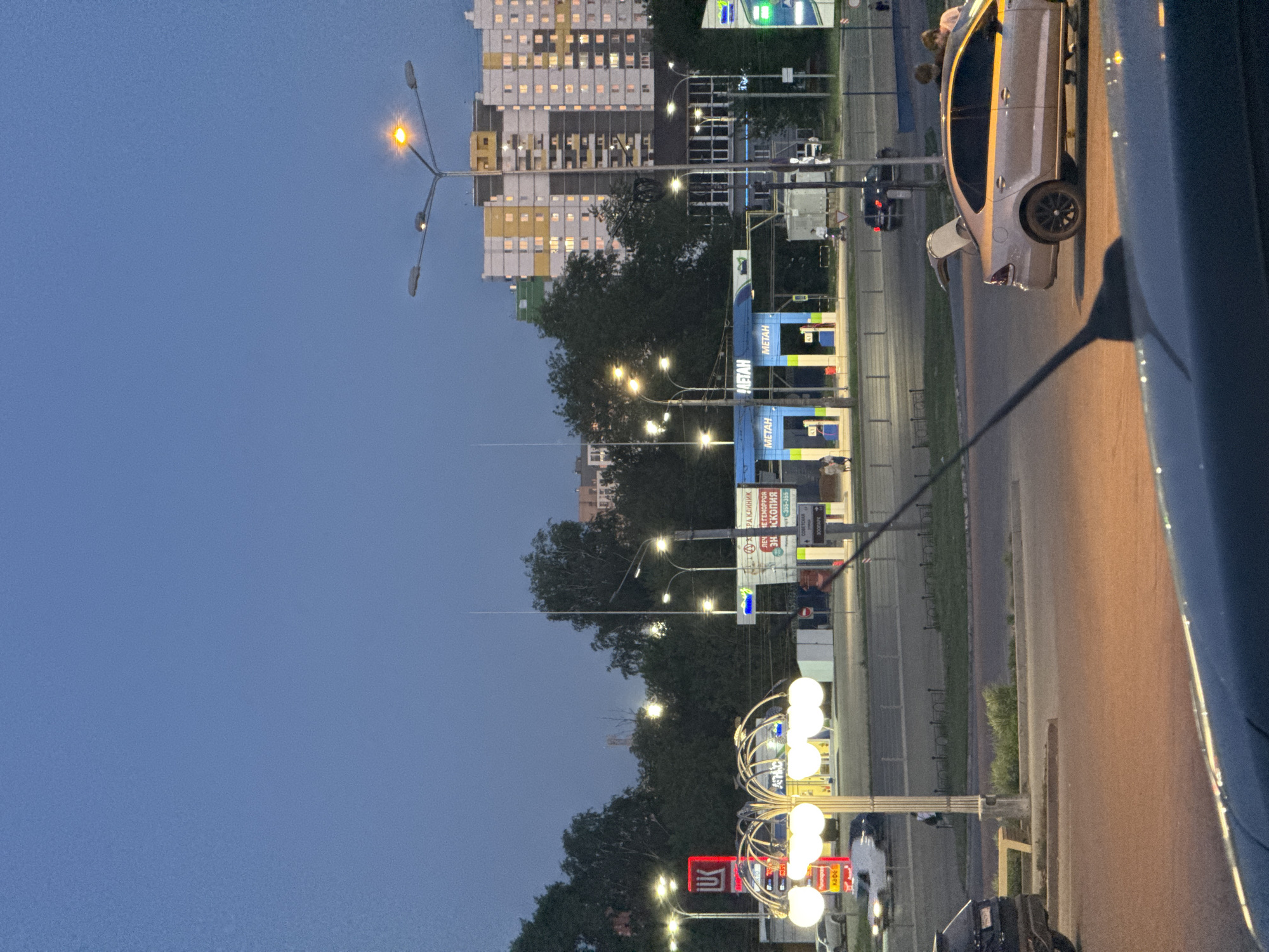 The image shows a street in Izhevsk, Russia, at dusk. Several lampposts illuminate the road, and a tall apartment building is visible in the background. A gas station and various commercial signs line the street. A light-colored car with an open back door is partially visible on the right. Another car is visible on the road. The sky is a deep blue, indicating the late hour.