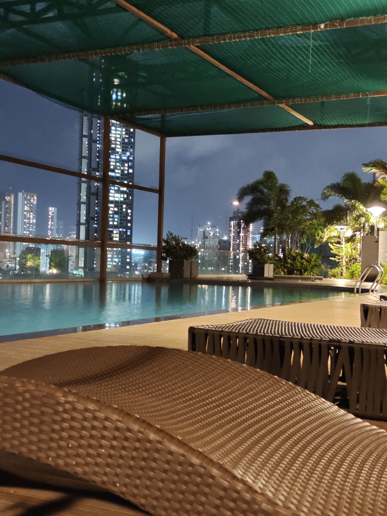 This image depicts a nighttime view of an infinity swimming pool on a rooftop in Mumbai, India. The pool overlooks a densely populated cityscape illuminated by numerous building lights. In the foreground, two lounge chairs made of woven material are visible, one in focus and the other blurred. Beyond the pool, the skyline features a cluster of tall skyscrapers, some with distinctive architectural designs and illuminated windows. Palm trees and other lush greenery are strategically placed around the pool area, suggesting a tropical or subtropical urban environment. Several outdoor lamps provide ambient lighting. The sky is dark, indicating it is nighttime, with some cloudy formations. The overall scene conveys a sense of urban luxury and relaxation amidst the vibrant backdrop of Mumbai's nighttime panorama.