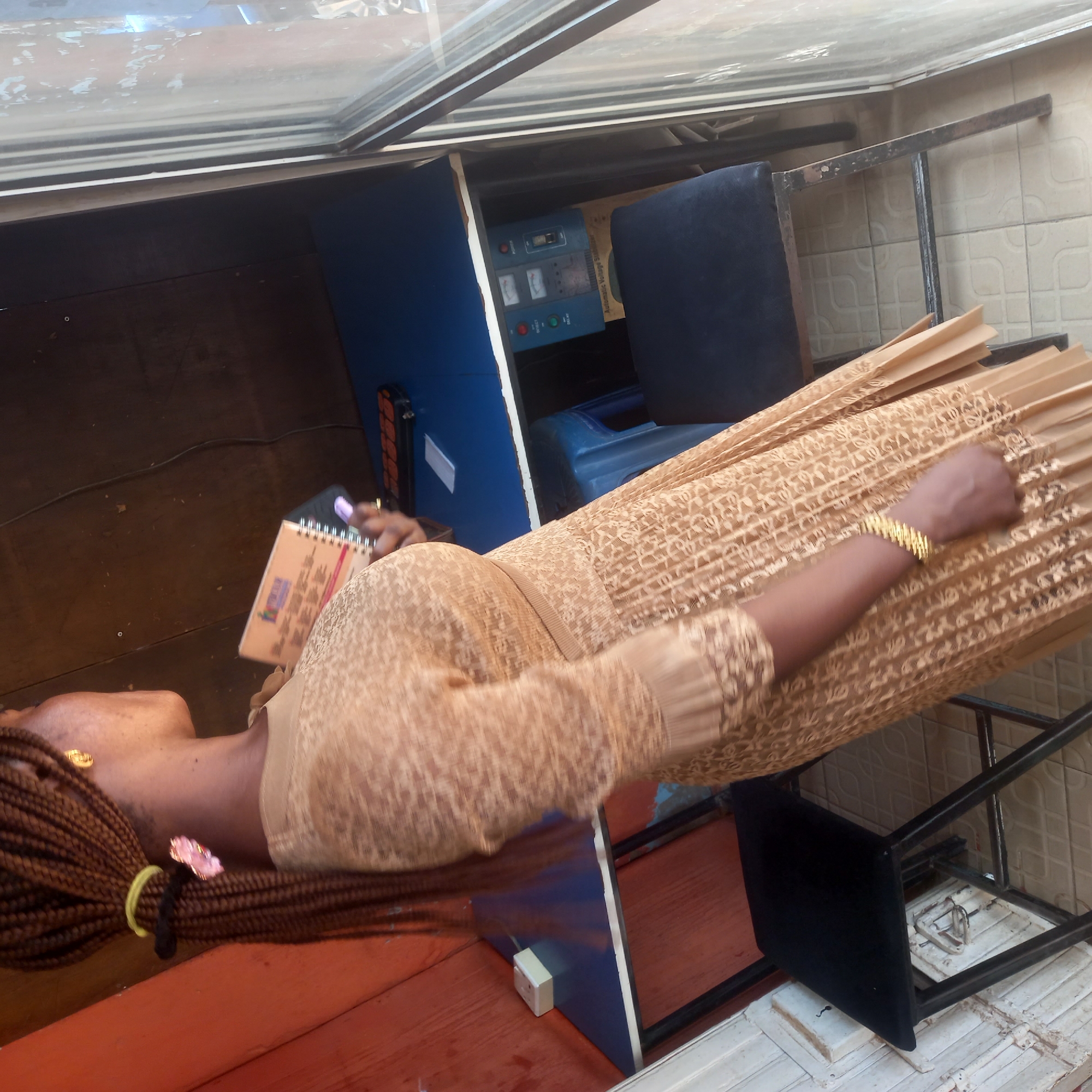 A person with dark skin and long, brown braided hair, secured with a yellow elastic and a pink flower clip, is depicted in an indoor or semi-outdoor setting. She wears a golden earring and a gold-colored chain-link bracelet on her left wrist. Her attire consists of a light brown, patterned dress or skirt with a matching top, featuring an intricate, lighter-toned design. She is holding a small, spiral-bound notebook with an orange/beige cover in her left hand, suggesting she is taking notes or reviewing information.
The background includes a dark brown floor and a light-colored tiled wall. Overhead, a glass or transparent canopy suggests a covered, sunlit space, indicating daytime. Behind the person, a blue and white utility stand holds an electronic device explicitly labeled "Automatic Voltage Stabilizer." This device features two analog gauges, multiple buttons, and indicator lights. A dark upholstered chair with a black metal frame is positioned nearby. Other objects visible include a black power strip or cable manager on the floor and a small white electrical box on the blue stand. The overall scene suggests a functional workspace, possibly an office or small business, typical for locations where power stability equipment like a voltage stabilizer is commonly used, such as Dadi Riba, Nigeria.