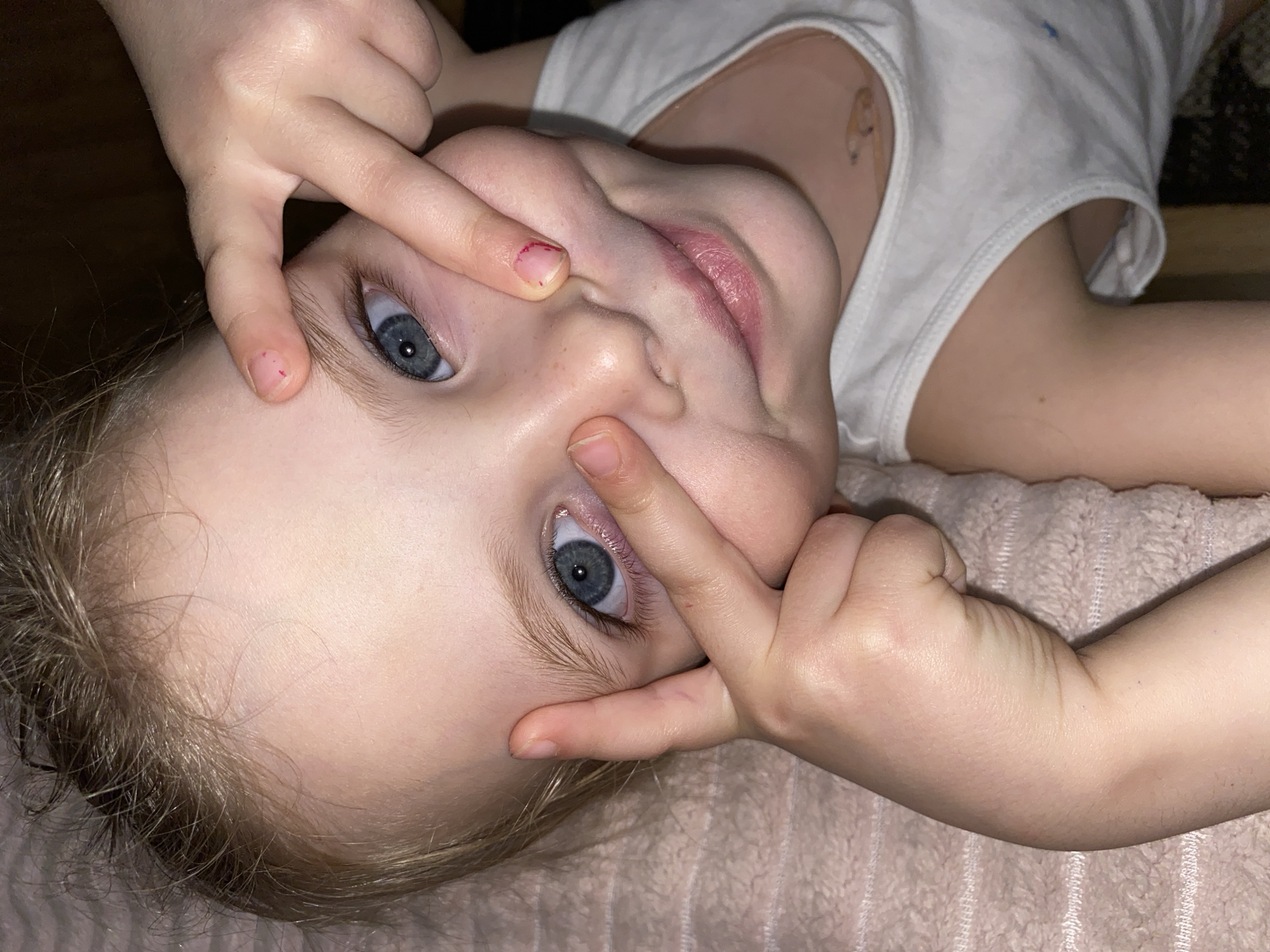 A close-up, rotated portrait captures a young child lying down indoors in Votkinsk, Russia, engaging in playful facial contortions. The child, with fair skin, light brown hair, and striking blue eyes, looks directly at the camera with a mischievous grin. Their fingers are pressed against their face, pulling at the skin around their eyes and nose, creating a humorous expression. The child is wearing a simple white sleeveless top or undershirt. Small reddish marks are visible on their fingernails, suggesting recent play. The child rests on a soft, textured, light pinkish-beige blanket or towel. The background is dark and indistinct, consistent with an indoor setting under soft lighting. No text is visible in the image.