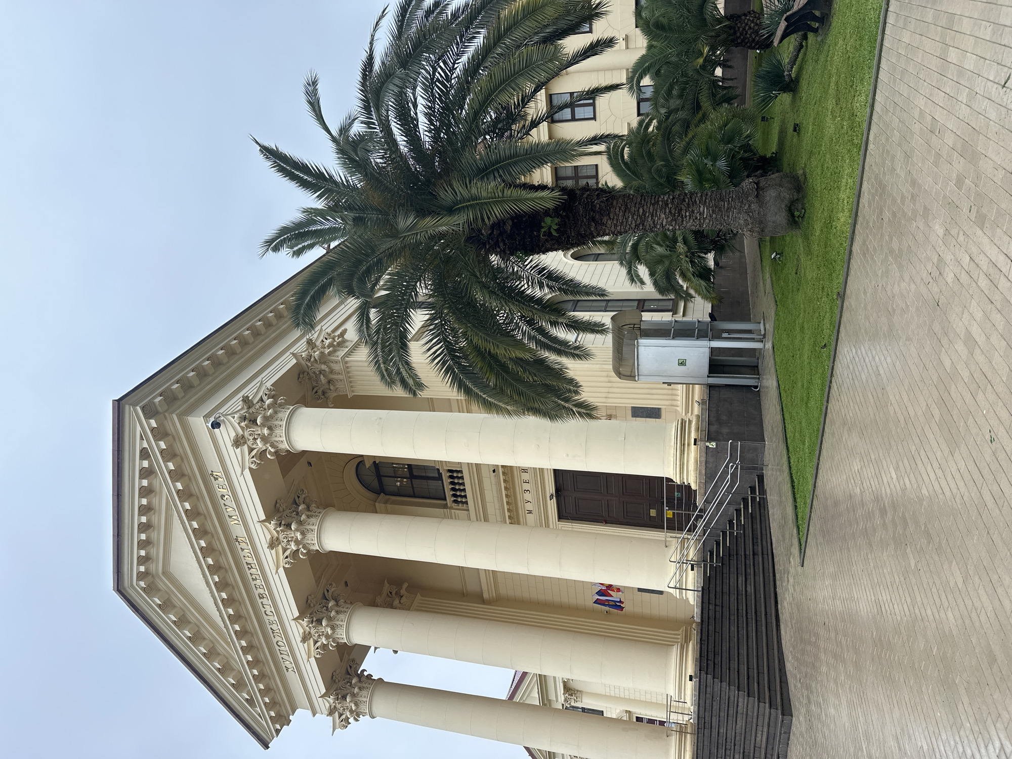 The image shows a low-angle view of the Sochi Art Museum, a neoclassical building with a prominent pediment and large columns. The museum is located in Sochi, Russia. A tall palm tree stands in front of the building, partially obscuring its facade. A patch of green grass is visible on the right side of the image, next to a paved walkway. A wheelchair lift is located on the left side of the building. The sky is overcast.