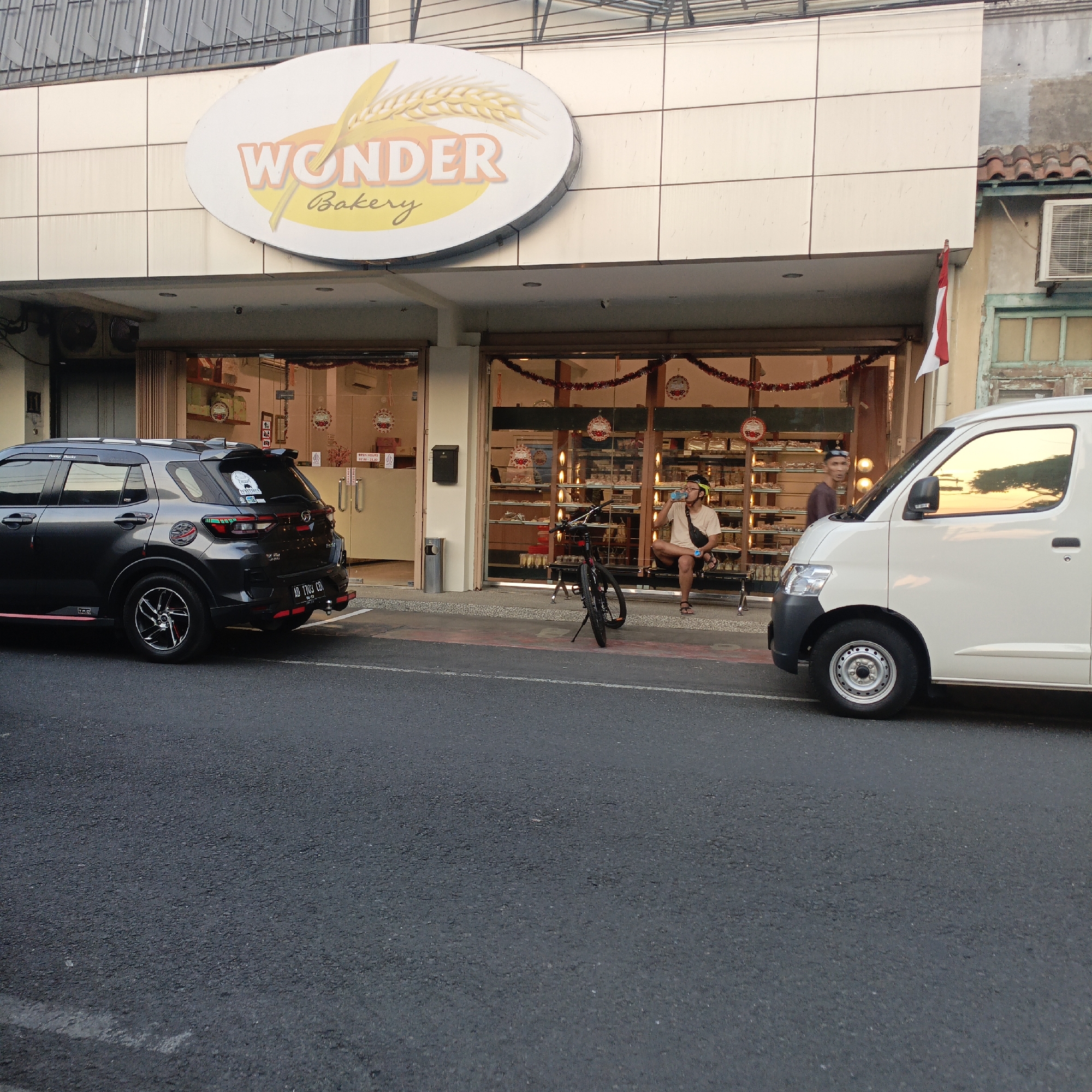 The image captures a street scene in Salatiga, Indonesia, featuring a "Wonder Bakery" storefront. A dark-colored SUV is parked on the left, and a white van is on the right. In front of the bakery, a person wearing a helmet and light-colored t-shirt is sitting on a bench, drinking from a bottle. A bicycle is parked nearby. The storefront displays baked goods behind glass, and festive decorations, including red garland, are visible. The overall ambiance suggests a commercial area in a city.