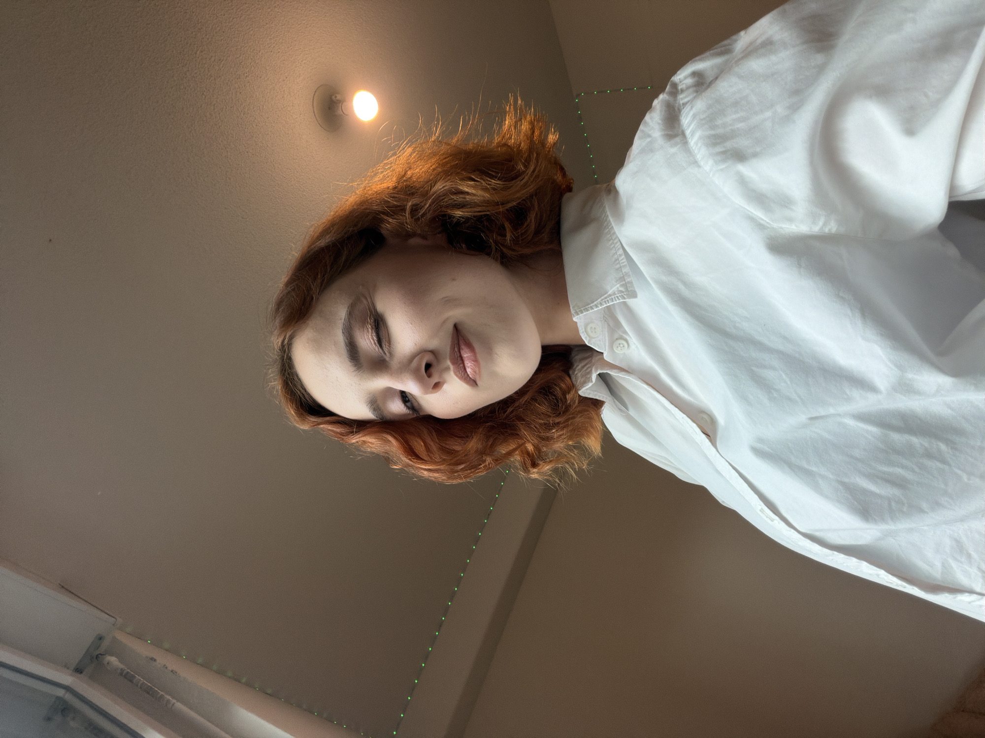 A young woman with auburn, wavy, shoulder-length hair and fair skin is captured in a low-angle shot, looking directly at the camera with a slight wink. She is wearing a crisp white collared shirt, the top buttons undone, revealing the collarbone. Her makeup includes subtle eyeshadow and lip color. The background is an indoor setting with a neutral-toned wall and ceiling. A single, bare lightbulb is visible on the ceiling, casting a warm glow. A string of small green lights runs along the top edge of the wall on the left side of the frame. The overall lighting suggests an indoor environment, possibly a room in a residential building. There is no identifiable indication of the location being Strasbourg, France.