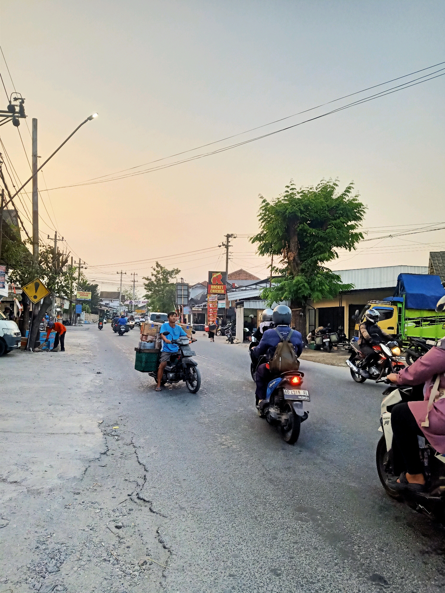 The image depicts a street scene in Klaten, Indonesia, likely during late afternoon given the warm, hazy light. The street is paved with cracked and weathered asphalt, with a drainage ditch running along the right side. Overhead, electrical wires crisscross the sky, supported by utility poles.
Several individuals are present, predominantly riding motorcycles. In the foreground, a person on a motorcycle is transporting a large cargo of boxes and green baskets. To their right, another person on a motorcycle, wearing a helmet, is riding ahead. Further down the street, a group of motorcyclists are visible, some carrying passengers. One motorcycle has a license plate starting with "AD 4319 XL".
On the left side of the street, businesses with signage are visible, including one that appears to be a restaurant with a "Rocket Chicken" sign. A person in an orange shirt is seen sweeping the sidewalk. Vehicles, including cars and a yellow truck with a blue tarp, are parked or moving along the street. A green tree with dense foliage stands prominently near the center of the image, casting a shadow on the street. The overall atmosphere suggests a bustling, everyday street scene in a commercial or mixed-use area of Klaten.