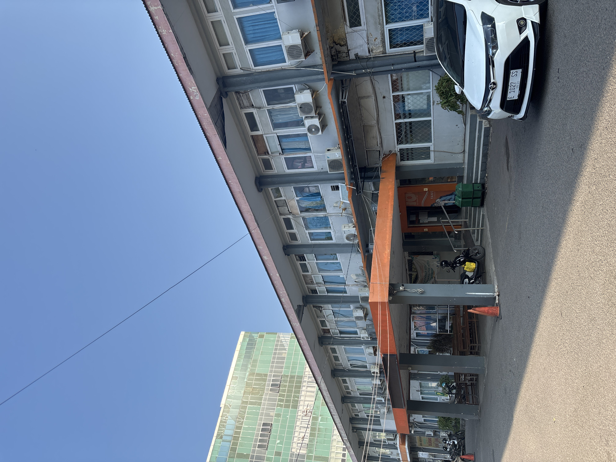 The image shows the exterior of a building in Cirebon, Indonesia. The building appears to be an institutional or office building with multiple stories and a grey facade. There are numerous windows, many with blue curtains visible, and air conditioning units are mounted on the exterior walls. An orange overhang extends across the front of the building, supported by dark grey columns. Parked in front of the building is a white sedan with license plate "E 1027 SS" and a black motorcycle. In the background, a taller building with a green-tinted glass facade is visible. The sky is clear and blue. The ground is asphalt, and there are some traffic cones present. The overall impression is of a functional, somewhat dated urban environment.