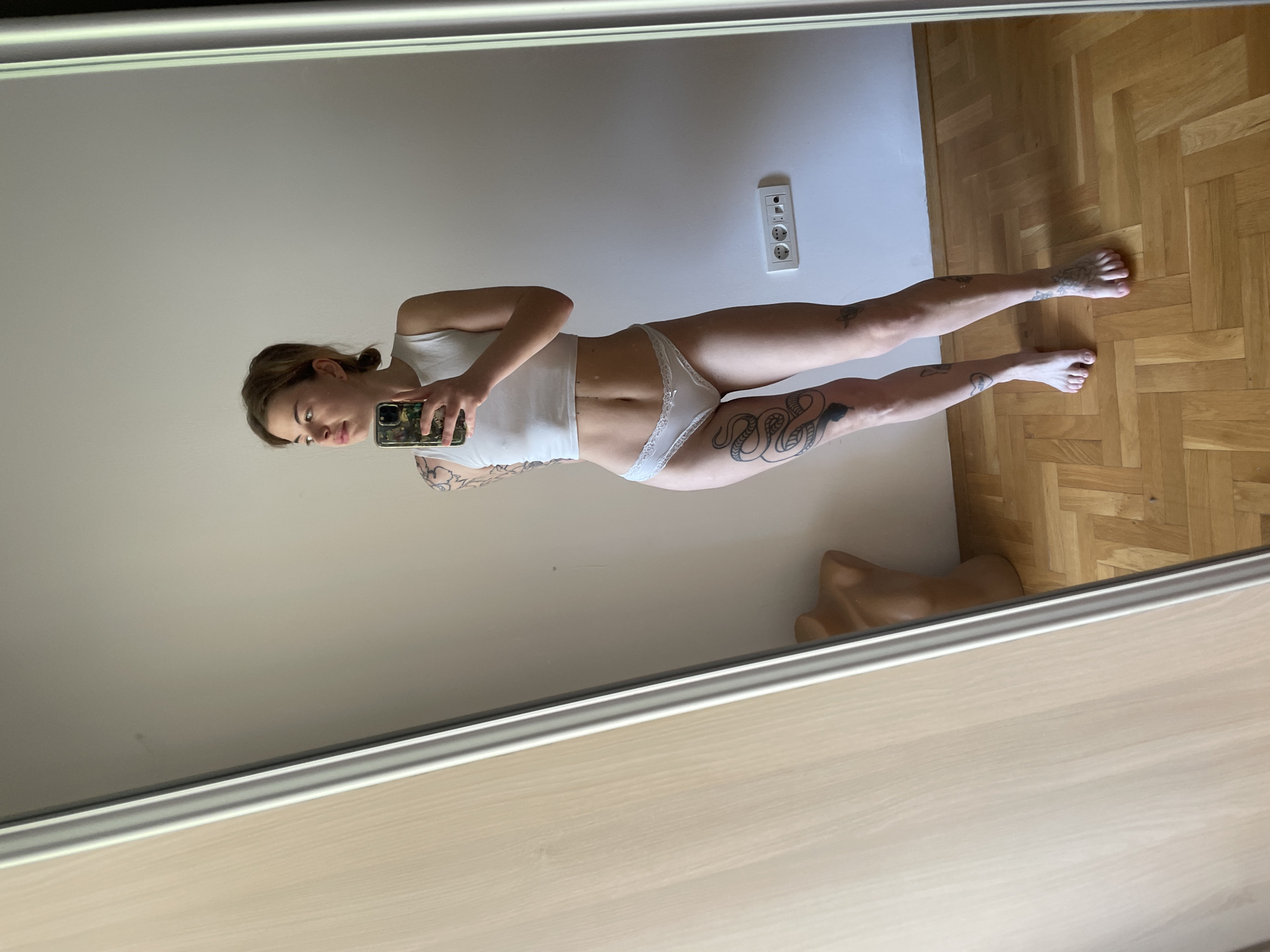 A full-length image captured in a mirror reveals a woman with fair skin and light brown hair pulled back in a loose ponytail. She holds a smartphone in her left hand, angled slightly downward as if taking a selfie. She is wearing a white tank top and white lace underwear. Tattoos are visible on her left arm and leg. She stands against a light beige wall. An electrical outlet is mounted on the wall behind her. The floor is made of wood planks arranged in a herringbone pattern. To the lower right of the woman, just within the mirror's reflection, is what appears to be a mannequin torso made of tan plastic. This photograph does not appear to be taken in Boreti, Montenegro.