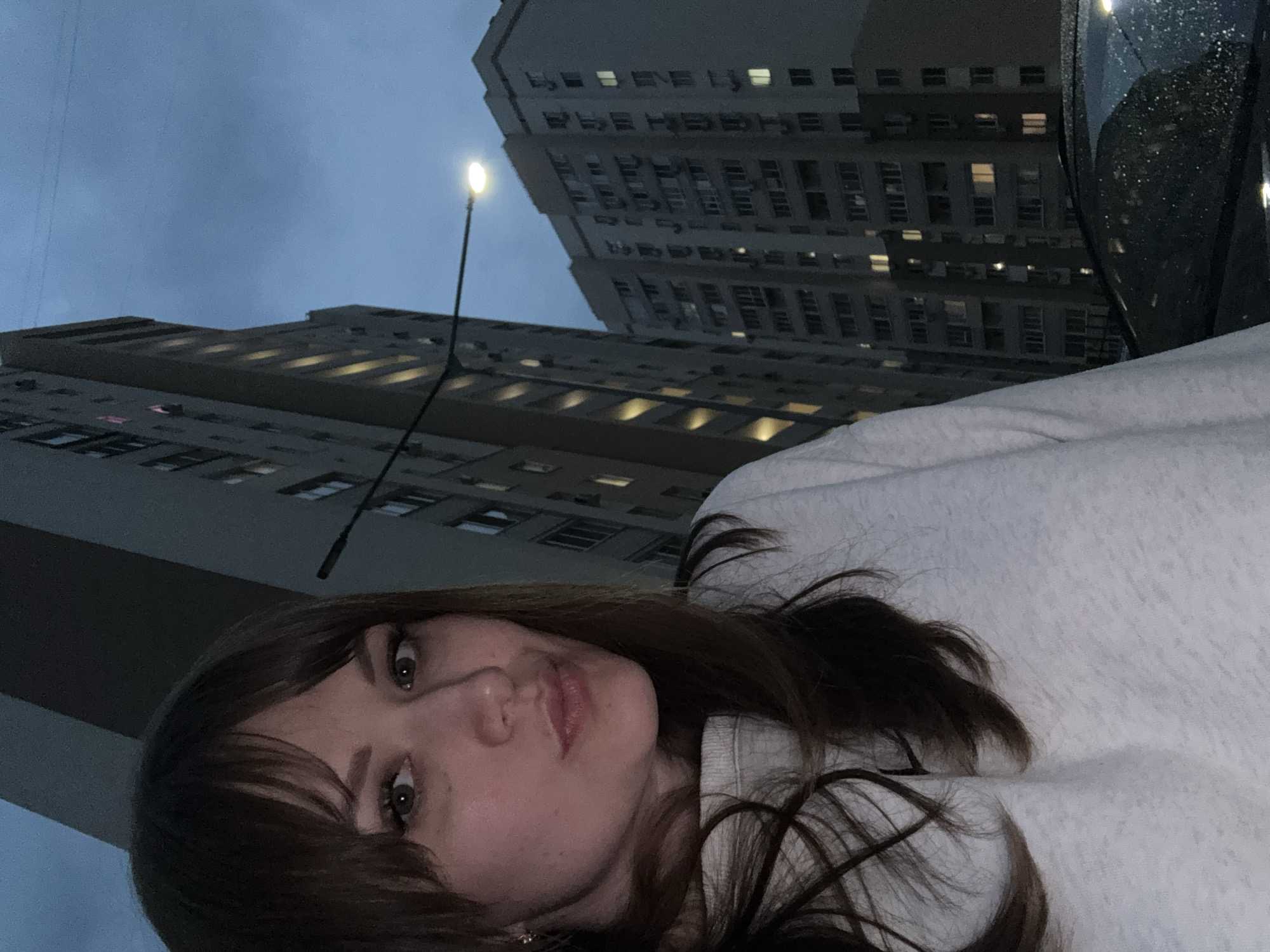 A woman with dark hair and fair skin is positioned in the foreground, her face angled toward the camera. She is wearing a light-colored sweatshirt. Behind her, towering buildings dominate the background, suggesting an urban setting. A street lamp is visible between the buildings. Based on the buildings' architecture and the background elements, the location is likely in Saratov, Russia. A portion of a vehicle is visible in the upper right corner of the frame. The sky is overcast.
