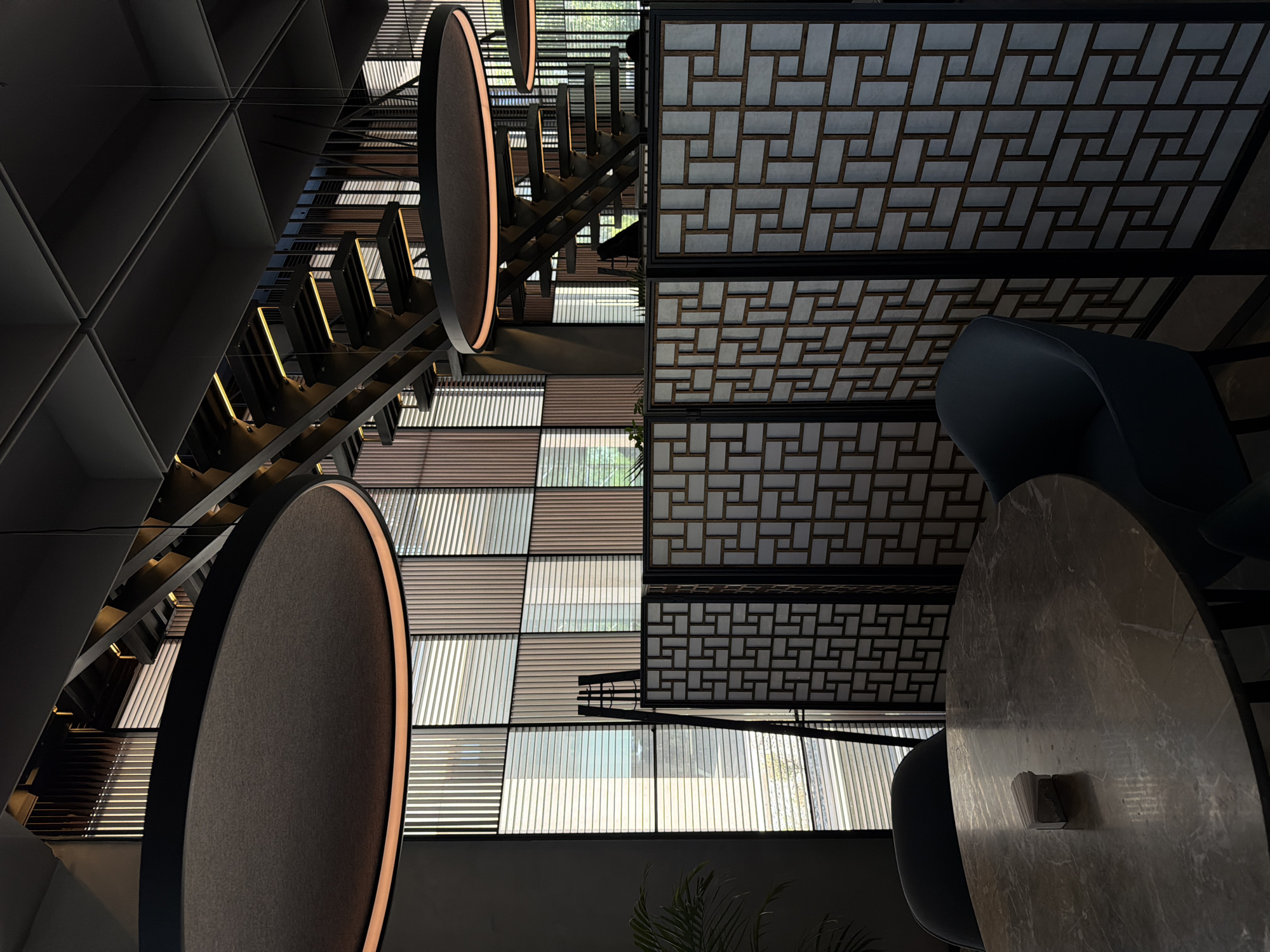 This is an interior shot taken in Bishkek City, Kyrgyzstan. The composition is oriented vertically and primarily features architectural and interior design elements. A staircase with metal supports and integrated lighting is visible on the left side of the image. Hanging circular light fixtures are prominent, one in the foreground and another in the background. Windows featuring a pattern of horizontal blinds or slats are central to the composition. A decorative ceiling with a geometric grid pattern is positioned above a dining area composed of a round table and two dark chairs. The table appears to be made of marble, displaying a light-colored grain pattern.